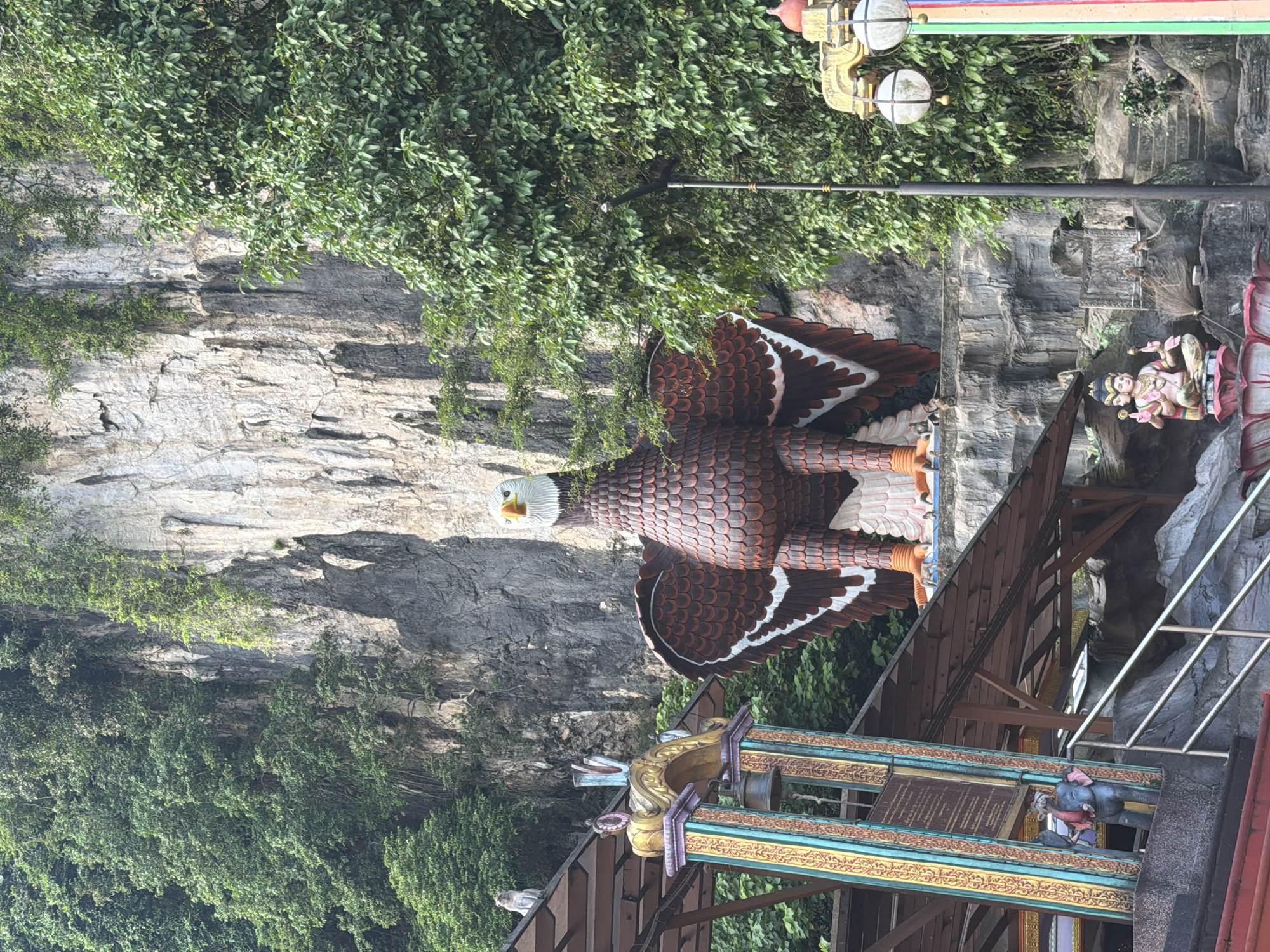 This image captures a scene at the Batu Caves temple complex, located in Gombak, Selangor, within the Selayang Municipal Council area of Malaysia. In the foreground, a large, detailed sculpture of a brown and white eagle stands prominently on a rocky ledge. The eagle sculpture has its wings spread, showcasing intricate feather details and bright orange feet.
To the left of the eagle, a colorful entrance structure is visible, adorned with gold, blue, and red painted pillars and an arched gateway. A bell hangs from the gateway. Further to the left, on the roof of a covered walkway, a small monkey is perched. Below this structure, a statue of a multi-armed figure seated on a lotus flower is visible, with another small statue of an elephant to its left.
The background is dominated by a massive, sheer limestone cliff face, partially covered by lush green foliage. Several smaller monkeys can be seen scattered on the rocky ledges and steps leading up the cliff. To the right of the eagle sculpture, a tall black pole with two white, spherical light fixtures extends upwards. The overall environment suggests an outdoor religious or cultural site nestled against a natural rock formation.
