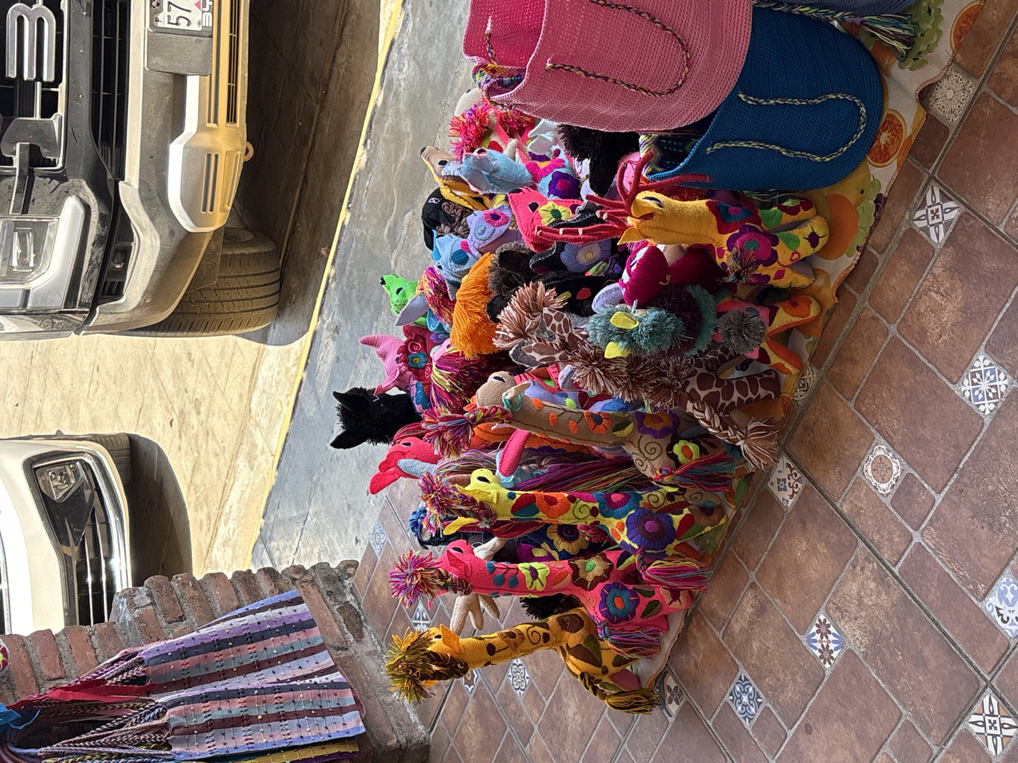 A roadside vendor's display in Puerto Escondido, Mexico, features an array of colorful, handcrafted stuffed animal toys, primarily giraffes, and bags. The toys, made of patterned fabrics and adorned with tassels, are piled atop a table covered with a printed tablecloth. Beside the display, two vehicles are visible: a white sedan and a black truck, partially obscuring the scene. The vendor's setup is situated on a tiled area. Nearby, a brick wall supports woven blankets.