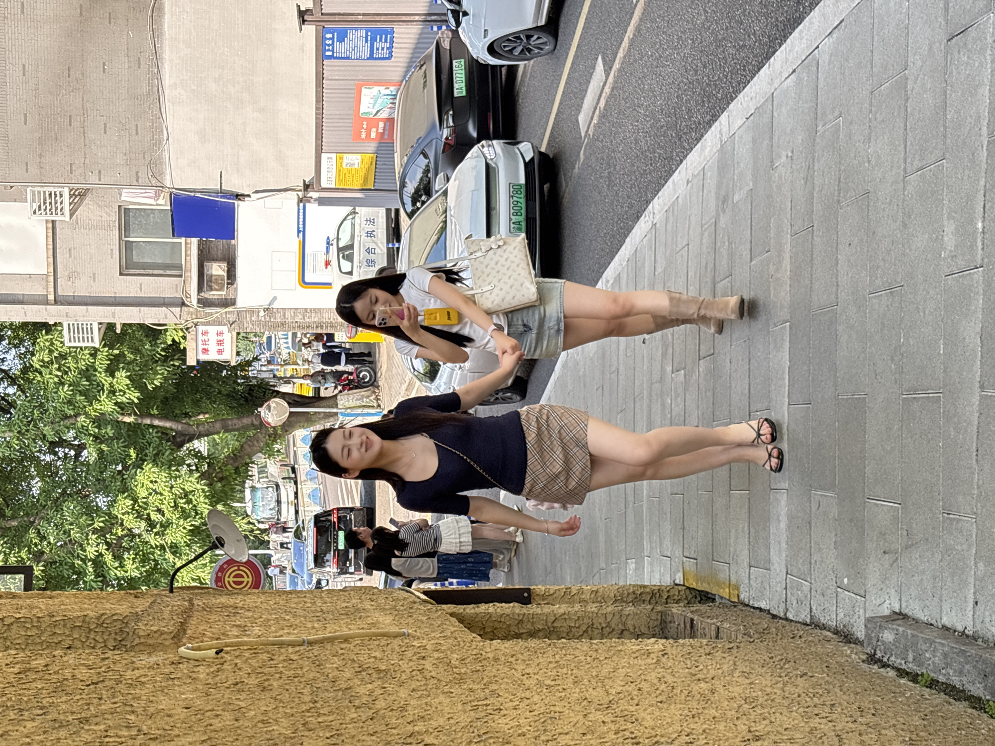 A full outdoor shot captures two young women strolling along a sidewalk adjacent to a street in Yuzhong District, China. The woman in the foreground wears a dark, short-sleeved top and a tan plaid mini skirt, paired with black sandals. The other woman is wearing a white top, a denim skirt, and tan boots; she is using a phone. Both have long dark hair.
The scene is set on a sidewalk made of gray pavers, with a street running beside it. Cars and scooters are visible in the street, along with the green license plates with white lettering on the vehicles.
A yellow building and a tree are present on the left side of the frame.