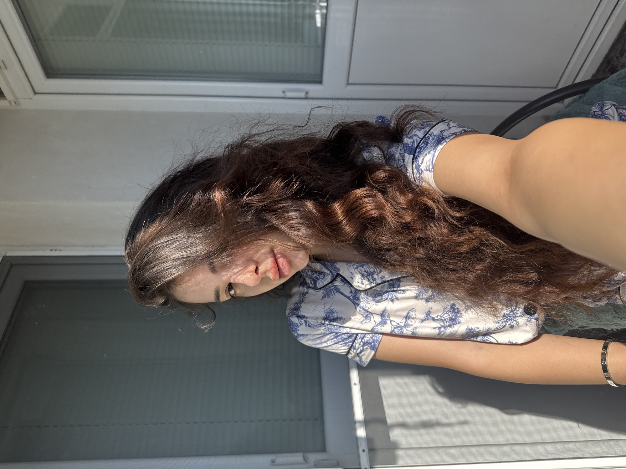 A young woman with long, dark, wavy hair is pictured outdoors, possibly on a balcony, in Plovdiv, Bulgaria. She is wearing a white and blue patterned short-sleeved pajama top. Her right arm is extended towards the camera, as if taking a selfie. She is wearing a silver bracelet on her left wrist. The background features a window with blinds and a white door frame, with strong sunlight casting shadows.