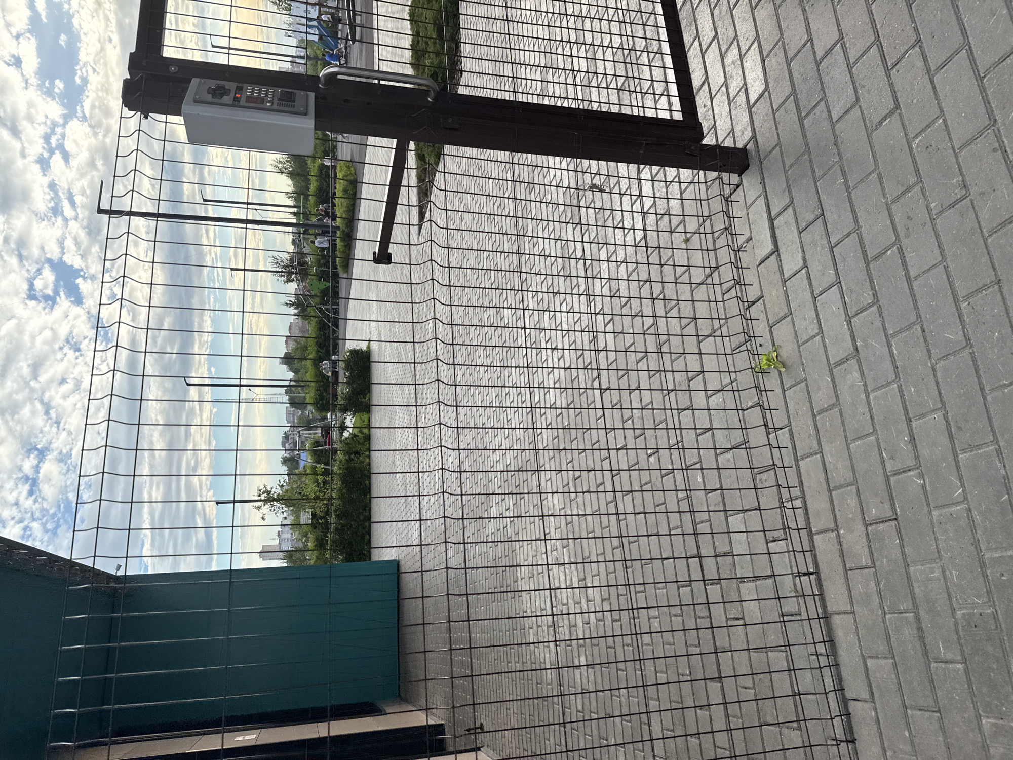 An open gate with a metal mesh fence stands in the foreground, leading to a paved area and a playground. A grey keypad with red and white buttons is mounted on the gatepost. Beyond the gate, a playground with swings and slides is visible, with a few people scattered throughout. The sky is partly cloudy, and buildings can be seen in the distance. The paving stones are grey and rectangular. This scene is consistent with a public park or residential complex in Perm, Russia.