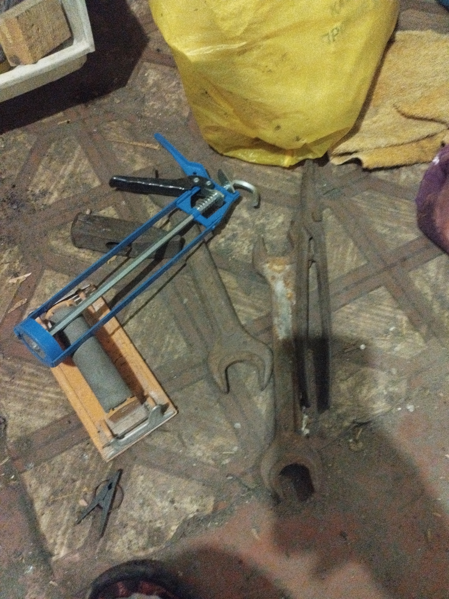 The image displays a cluttered ground surface, possibly a workshop floor, with various tools and materials. In the foreground, a blue caulking gun is prominently positioned. To its right, several large, rusty wrenches are laid out. A yellow plastic bag is visible in the upper right portion of the image, partially obscuring a tan fabric. A small black clamp lies near the bottom left. The ground is a textured surface with visible lines and debris. The overall scene suggests a working environment where tools are in use or stored. The location is specified as the city of Sarykol, Kazakhstan.