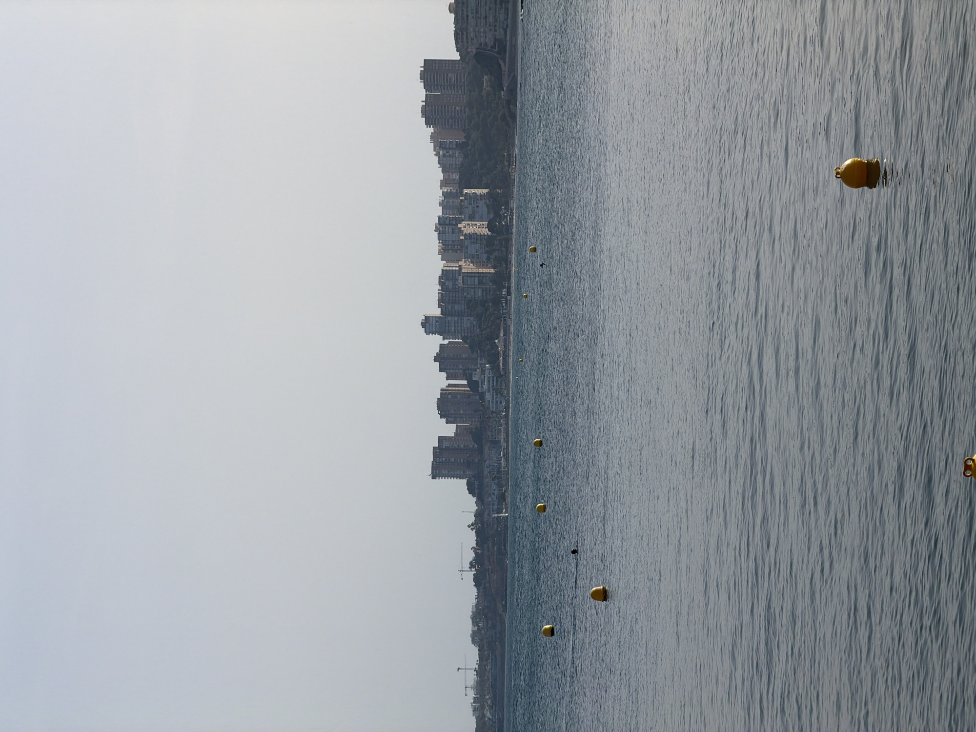 The image shows a wide shot of a calm body of water with a city skyline in the distance. Several yellow buoys are floating on the water's surface. In the middle ground, a person is partially submerged in the water, appearing to swim. The city skyline consists of numerous tall buildings, some of which are residential high-rises. Construction cranes are visible on the far left of the skyline, suggesting ongoing development. The sky is overcast, creating a soft, diffused light. The water is a muted blue-grey, reflecting the subdued lighting conditions. The overall scene conveys a sense of a coastal urban environment on a hazy day.