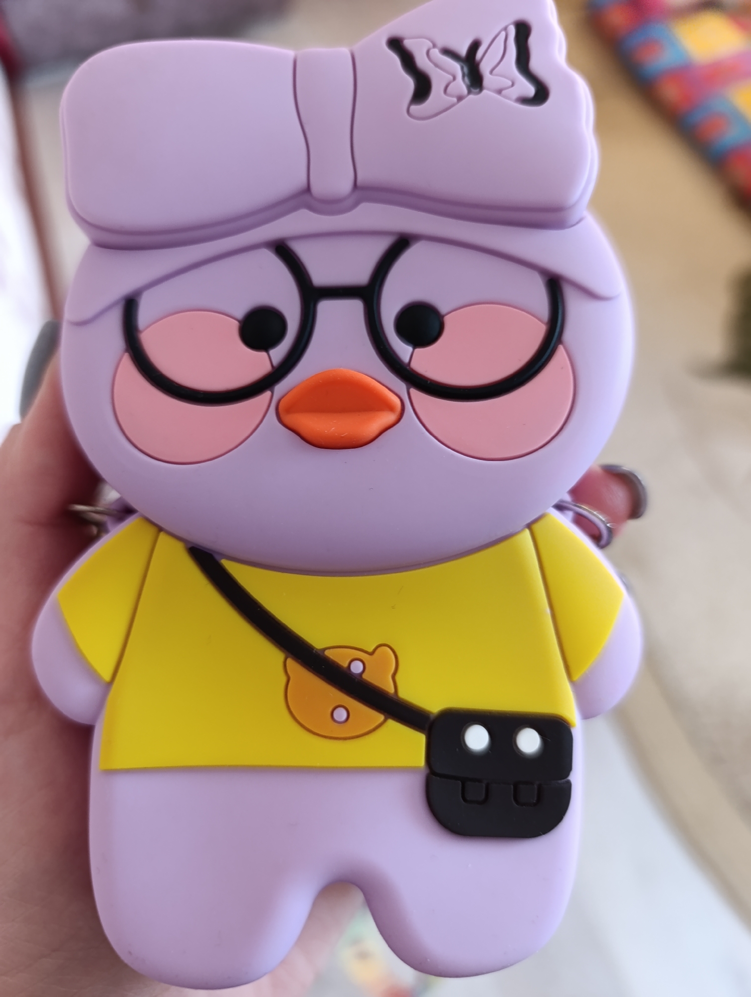 A close-up shot of a cartoon-style keychain featuring a purple, stylized figure wearing glasses and a yellow shirt with a satchel. The figure has a bow on its head. The keychain is held in a person's hand. The specific location is not discernible from the image.