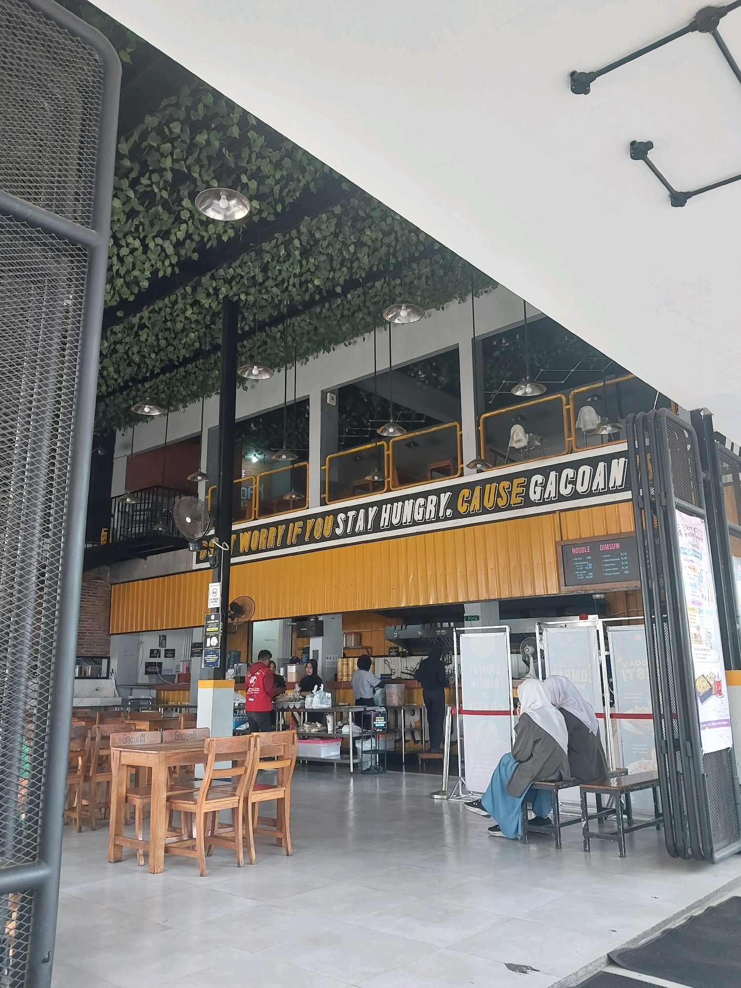 The image captures the interior of a restaurant with an industrial and modern design. The ceiling is adorned with artificial greenery and illuminated by hanging pendant lights. The walls of the main dining area are painted yellow, featuring prominent text that reads "DON'T WORRY IF YOU STAY HUNGRY, CAUSE GACOAN". This phrase is a slogan for the restaurant chain "Gacoan" which is known for its spicy noodles and is popular in Malang, Indonesia.
In the foreground, several wooden tables and chairs are arranged, suggesting a seating area. A metal grate structure is visible on the left side of the image, partially obscuring the view. In the background, there are multiple levels within the restaurant, with balconies and seating areas on the upper floors. Staff members are visible behind a counter, preparing food or serving customers. Two women wearing hijabs are seated on a bench in the middle ground, facing away from the camera, appearing to be customers. The overall ambiance suggests a busy and casual dining establishment.