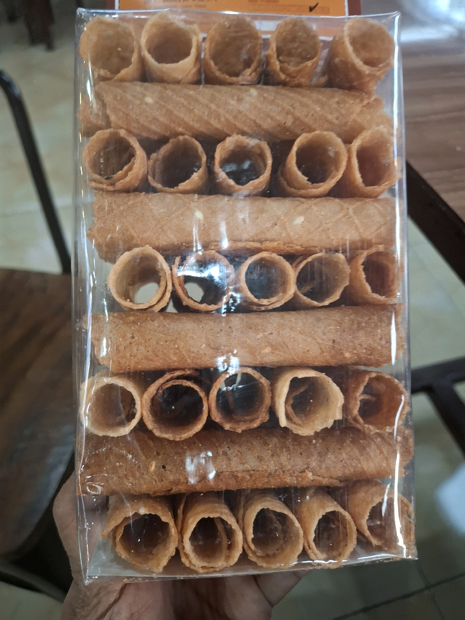 In Purworejo, Indonesia, a package of crispy rolled wafers, commonly known as *semprong*, is presented. The wafers are arranged vertically within a clear plastic container, showcasing rows of golden-brown, hollow cylinders. Some rows feature the rolled wafers standing upright, while others are laid horizontally, creating a layered effect. The packaging is partially obscured by a hand, suggesting the item is either being held or has just been picked up. The background hints at an indoor setting with furniture and a wooden surface, typical of a home or a small shop.