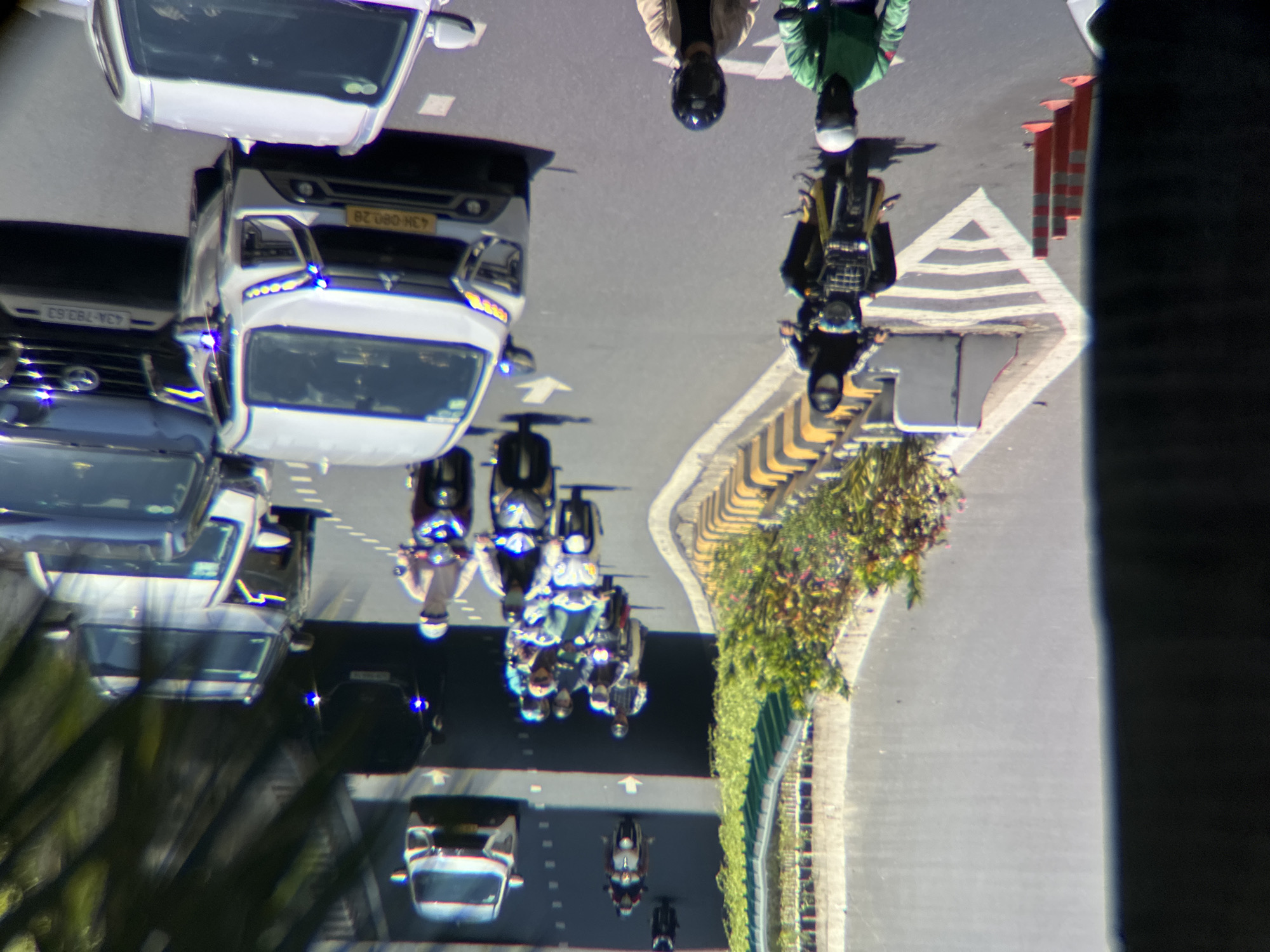 The image shows a street scene in Da Nang, Vietnam, with a traffic flow of cars and motorcycles. In the foreground, a person wearing a green jacket and helmet is riding a motorcycle. To their right, another person in a light brown jacket and helmet is also on a motorcycle. Further back, a white SUV is in the right lane, and a dark SUV is to its left. A motorcycle with a rider wearing a black outfit and mask is in the left lane, moving away from the viewer. Further up the road, a line of motorcycles and cars are visible, some in a tunnel or underpass. The road is asphalt, with white lane markings and traffic cones lining the side. Greenery is present on the left side of the road, with a low wall and yellow striped barriers. The scene appears to be during daylight, with shadows indicating sunlight.