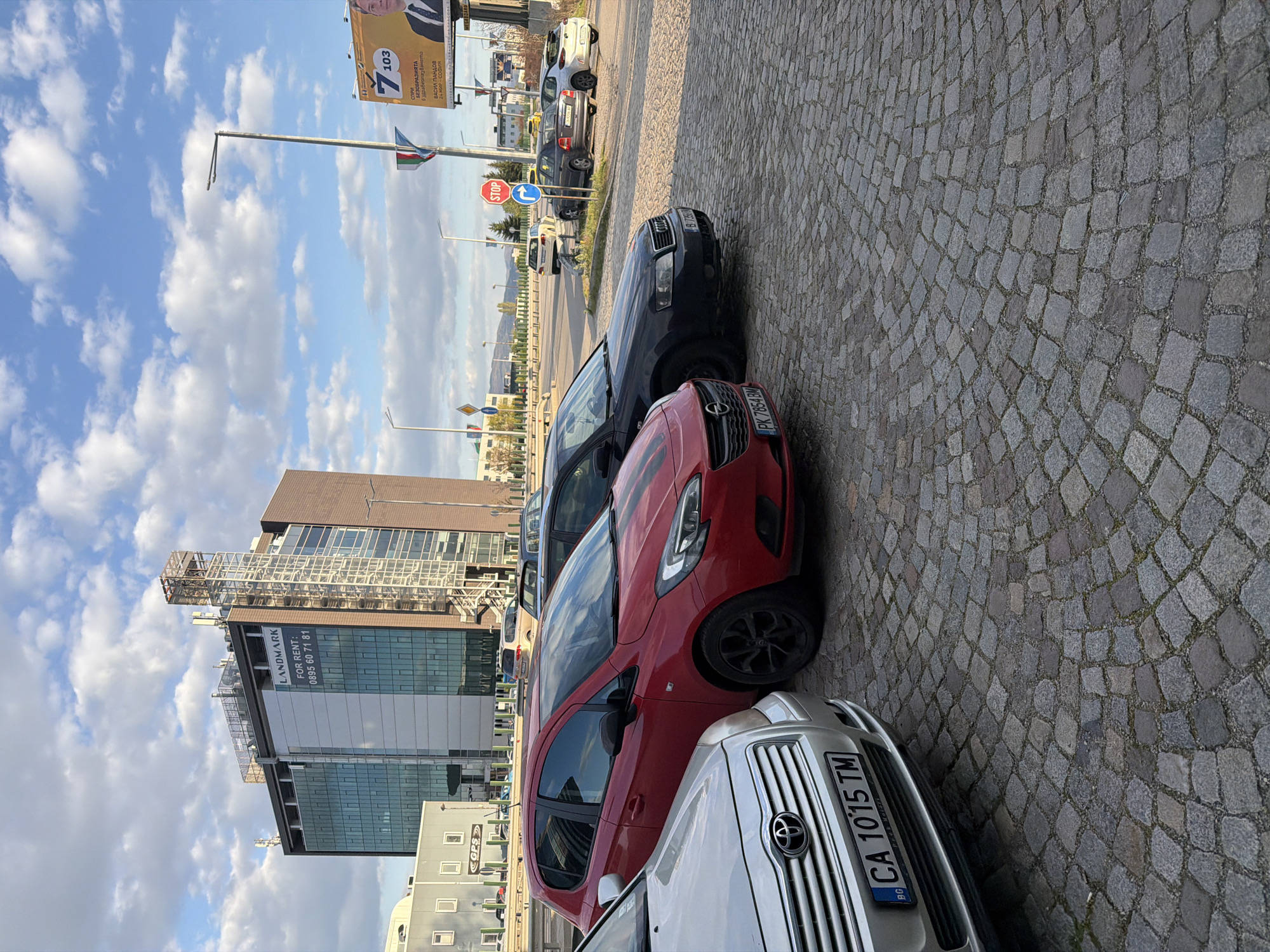 The image, originally captured in portrait orientation and rotated 90 degrees clockwise, displays a street scene in Sofia, Bulgaria, on a bright, partly cloudy day. The foreground features a cobblestone pavement alongside parked cars.
The primary subjects are several cars parked parallel to the street on the cobblestones. Closest to the viewer is the front of a silver Toyota, followed by the front-side of a red Opel Corsa, and then a dark grey or black Audi. Further down the street, more cars are visible, including a white SUV and other sedans.
In the midground, a street with light traffic is visible, lined with lampposts. Street signs, including a red "STOP" sign and a blue "turn right" arrow sign, are present. A large billboard dominates the midground sky, featuring a political advertisement.
The background shows modern urban architecture, including a prominent multi-story glass building with "LANDMARK" written on its side. Below this, text indicates "FOR RENT:" and a phone number "0895 60 71 81". Another smaller building in the distance has "CGPS" visible. The sky is blue with scattered white clouds, suggesting good weather conditions.
Visible text details include:
* **License plates**: "BG CA 1015 TM" (silver car, Sofia region), "BG PK 7854 BM" (red car, Plovdiv region), and "BG B7 7350" (dark car, Sofia region).
* **Billboard**: "7103", "Спри БЕЗОБРАЗИЯТА в здравеопазването" (Stop the outrages in healthcare), "ВАСИЛ ПАНДОВ" (Vasil Pandov), and "24 МИР - СОФИЯ" (24th constituency - Sofia).
* **Building**: "LANDMARK", "FOR RENT: 0895 60 71 81", and "CGPS".
* **Street sign**: "STOP".
The scene captures a typical urban environment with commercial buildings, parked vehicles, and street infrastructure, under natural daylight.