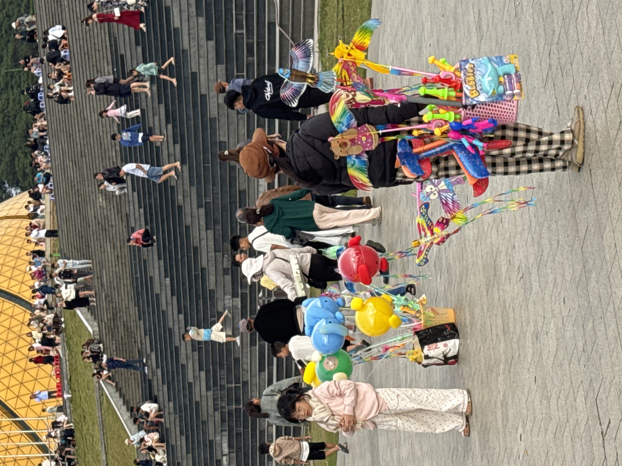 The image shows a busy outdoor scene with a large set of stairs in the background, filled with people. In the foreground, a street vendor is selling toys, primarily balloons and kites, from a basket. A young girl wearing pajamas and a scarf stands to the left of the vendor, looking at the camera. The location is identified as Da Lat, Vietnam, suggesting a tourist area. The overall atmosphere appears to be lively, with many people engaged in various activities.