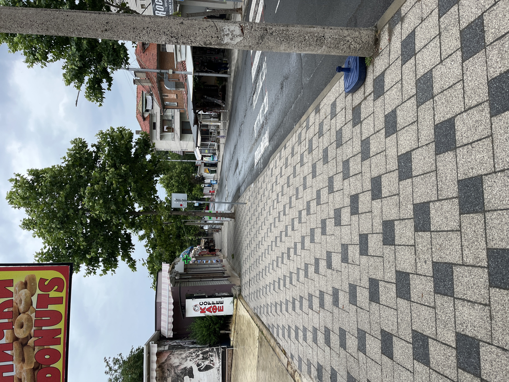 This is a street-level view of a sidewalk and road in Obzor, Bulgaria. The sidewalk is paved with interlocking rectangular tiles, alternating in a pattern of light gray and dark gray. A large concrete utility pole stands on the right side of the sidewalk, near a blue plastic base. The road, to the right of the sidewalk, appears wet and has a pedestrian crosswalk with white markings. Several small shops with awnings line the other side of the street. One shop has a sign with "DONUTS" written in large red letters. Another sign on a shop closer to the foreground reads "KAФE" (Coffee) in red letters. Trees with full green foliage are visible along the street. The sky is overcast. No people are clearly visible in the immediate foreground, but a few figures can be seen further down the street near the shops. The overall impression is of a commercial street scene on a cloudy day.