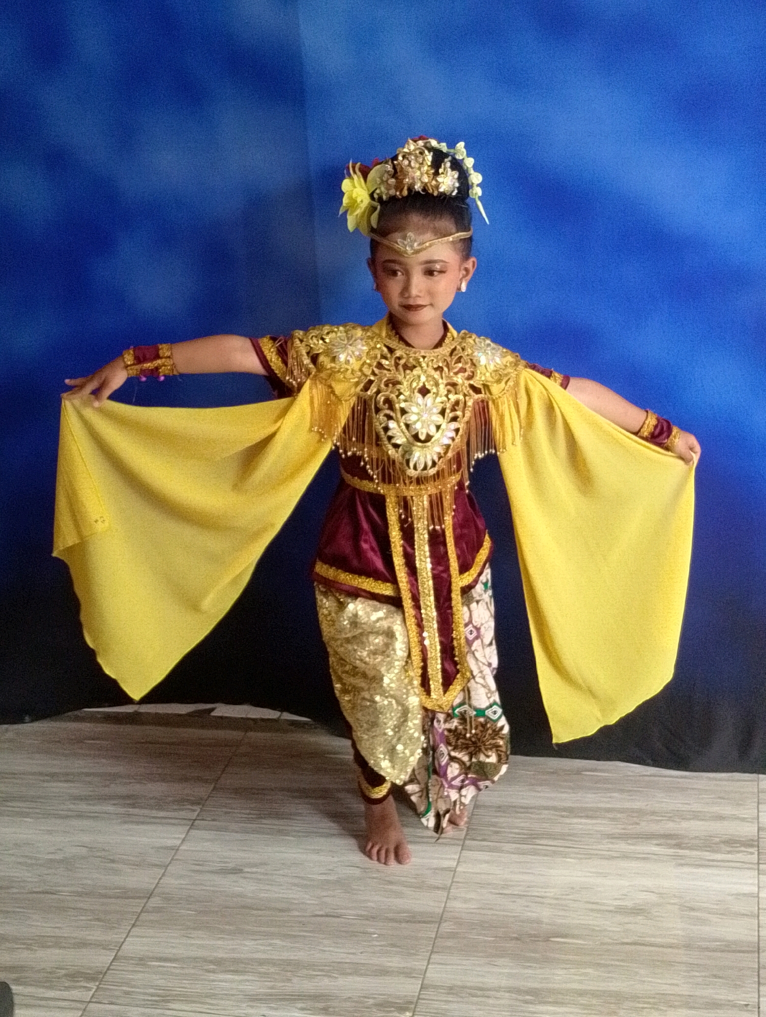 A young girl, likely of Indonesian descent, stands with her arms extended outwards, holding large, bright yellow fabric panels, in what appears to be a formal dance pose. She is adorned in a traditional, elaborate costume featuring a deep red velvet bodice with intricate gold embroidery and embellishments, including beaded fringe. Her lower garment consists of shimmering gold pants or leggings layered with a patterned sarong-like fabric. Her hair is styled up with an ornate headpiece adorned with flowers and gold accents, and she wears matching gold jewelry on her wrists. The background is a gradient blue curtain, with a dark fabric draped across the lower portion. The floor is tiled with a light gray, wood-grain pattern. The lighting suggests an indoor setting, possibly a studio or performance space. The image is taken from a slightly low angle, with the girl centered in the frame. The overall impression is of a cultural performance or presentation. The location of Gunung Putri, Indonesia, is noted for its batik and craft industries, which may explain the elaborate traditional attire.