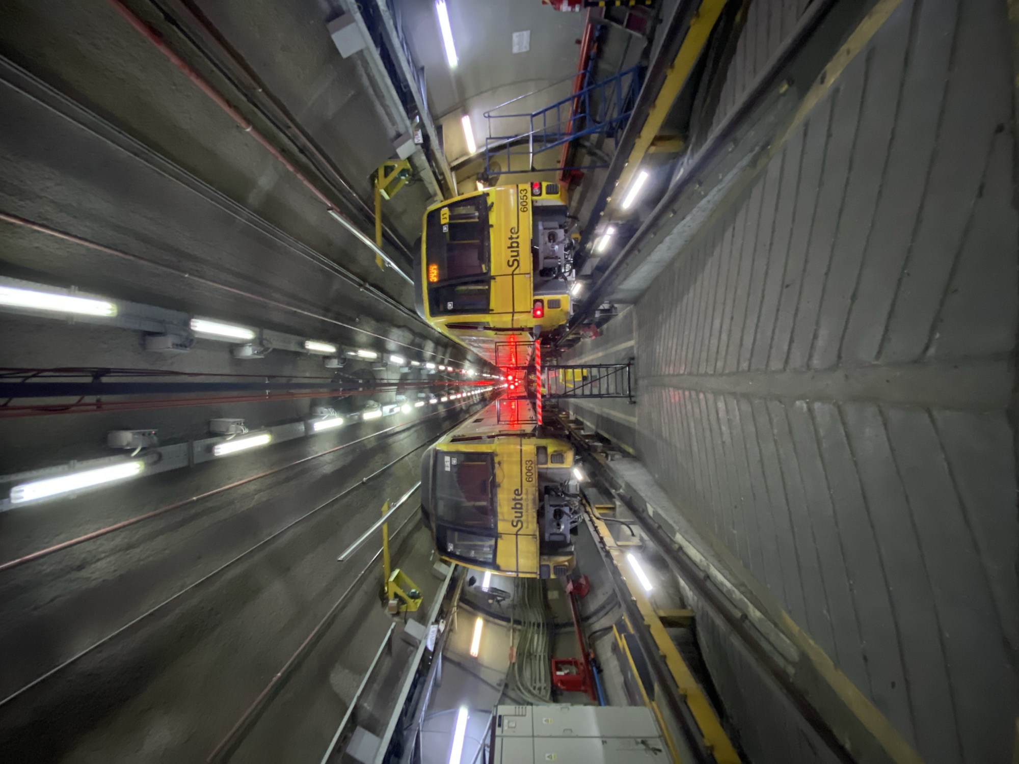 The image presents a highly symmetrical view of an underground tunnel, characteristic of a subway or train system, likely in Buenos Aires, Argentina. The composition is dominated by a single, bright yellow subway car, clearly visible in the upper half of the frame. Its distinct features include "Subte" and the identification number "6063" written on its side. A small number "11" is also visible on a side window.
A striking feature of the scene is the near-perfect vertical reflection that mirrors the subway car and the tunnel infrastructure in the lower half of the image. This reflection suggests the presence of a highly polished floor, water, or another reflective surface creating a symmetrical illusion, making it appear as if two identical trains are positioned head-to-head.
The tunnel itself is an industrial environment with concrete or gray-painted walls. Numerous long fluorescent light fixtures illuminate the tunnel, running along both upper sides and contributing to the bright, artificial lighting. Various pipes, conduits, and cables are visible along the upper walls. Around the upper subway car, yellow metal platforms with blue railings indicate a maintenance bay, depot, or workshop setting rather than an active passenger line.
In the far distance down the tunnel, a series of small, bright red lights create a distinct vanishing point, adding depth to the perspective. These red lights are also clearly reflected in the lower half of the image. White electrical boxes are visible on the side walls/floor. The overall atmosphere is stark and industrial, characterized by the consistent artificial lighting and the strong visual symmetry. The time of day is not discernible due to the underground setting.