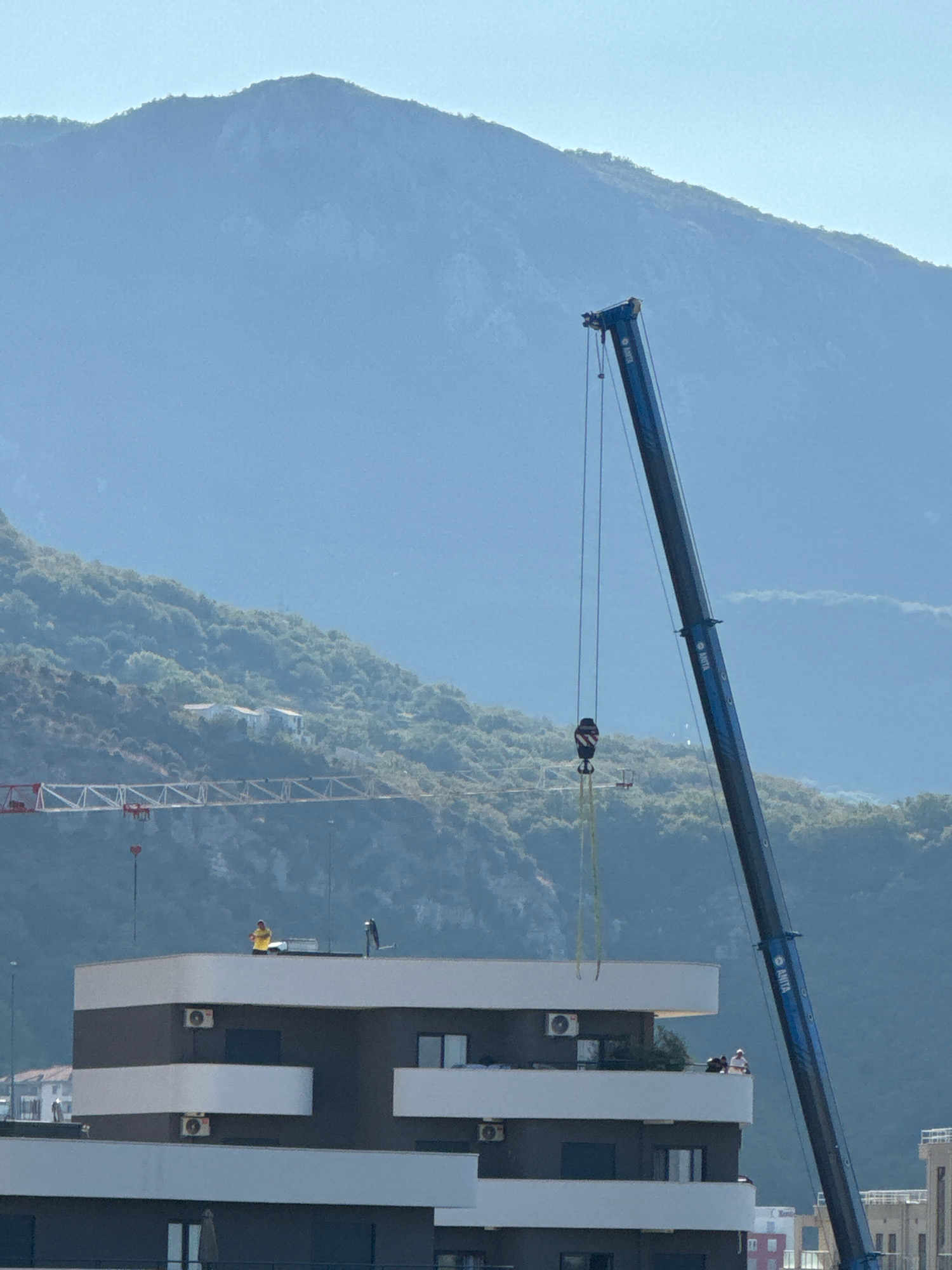 The image depicts a construction scene in Boreti, Montenegro, with a large blue crane dominating the right side of the frame. The crane's boom extends diagonally upwards and to the left, with its hook suspended by cables. In the middle ground, a modern building with dark and white horizontal bands is visible. Several people are present on the building's balconies and roof. One person in a yellow shirt stands on the roof. In the background, a misty mountain range rises, covered in dense green vegetation. To the left of the blue crane, a portion of a red tower crane is also visible, suggesting ongoing construction activity in the area. The sky is a clear, pale blue.