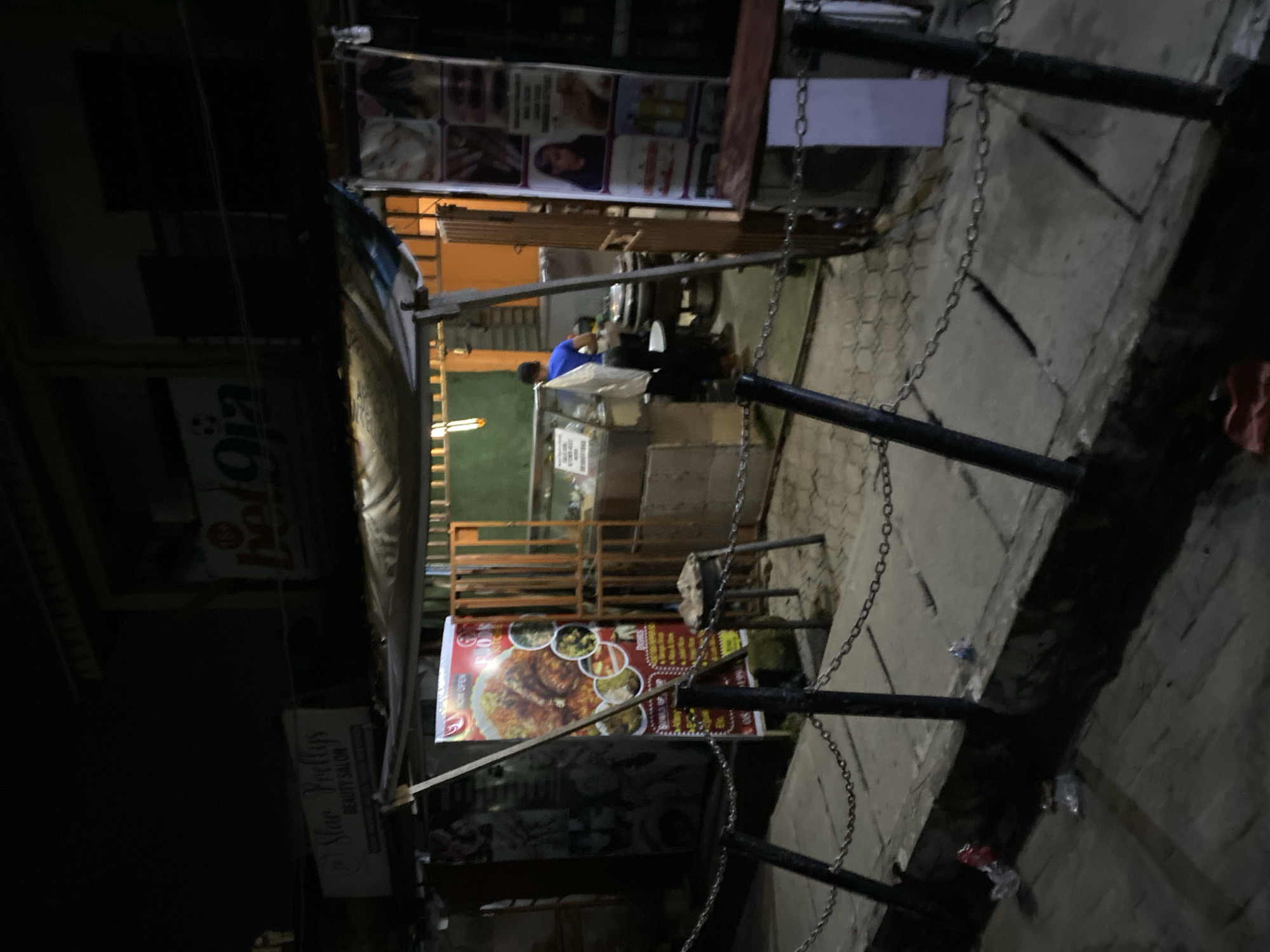 The image depicts a nighttime scene of a street in Ado, Nigeria, with a small food stall visible. The stall is illuminated by lights and has a sign displaying images of food items. Two individuals are present at the stall, one wearing a blue shirt. A sign for "Star Prettys Beauty Salon" is visible nearby. The street is paved and has a metal railing along the edge, with chains hanging from it. The environment is dark, suggesting it is late at night.