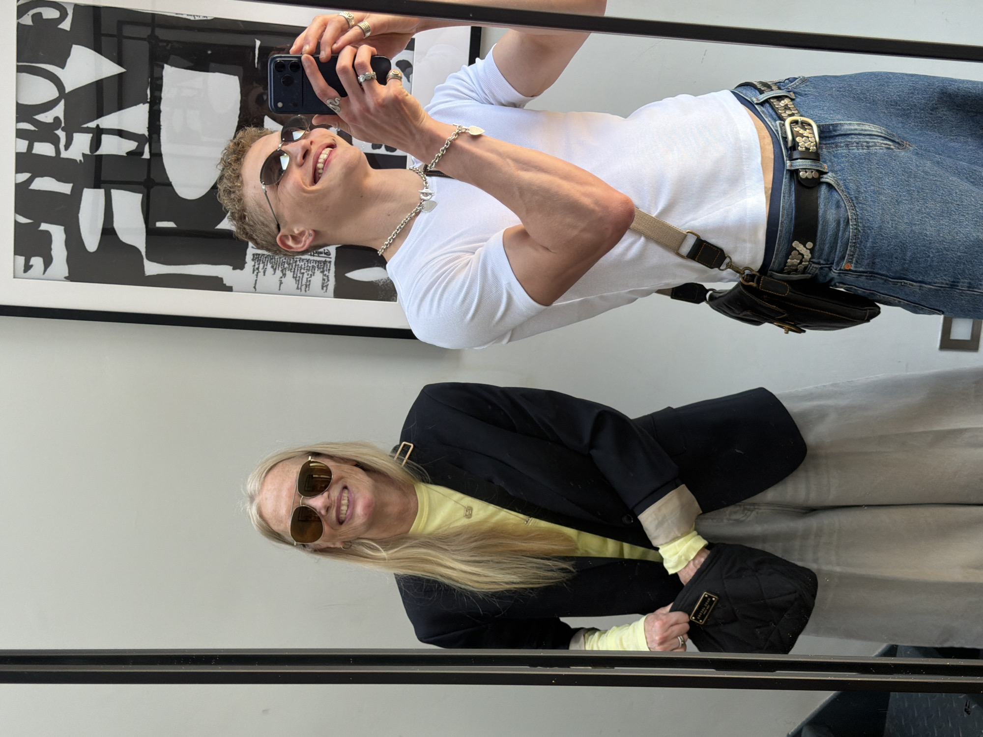 The image displays a mirror reflection of two individuals inside what appears to be a brightly lit indoor space, possibly in London, United Kingdom. The photo is taken from a perspective that captures the mirror's surface, showing both subjects looking towards the camera's reflection.
In the foreground, a young man with short, curly, light brown hair is taking a mirror selfie. He wears dark, round sunglasses and is smiling broadly, showing his teeth. His attire consists of a white ribbed short-sleeve t-shirt, blue denim jeans, and a black belt adorned with silver studs and an ornate silver buckle. He accessorizes with multiple silver rings on his fingers, a silver chain necklace featuring two heart-shaped pendants, and a matching silver chain bracelet. A small black pouch or fanny pack is attached to a light-colored cross-body strap. He holds a dark-colored smartphone with both hands, positioned to capture the image. Behind him, reflected in the mirror, is a large framed abstract black and white art piece with bold graphic shapes and some stylized text, where the letters "OVE" are partially visible.
In the background of the reflection stands an older woman with long, straight blonde hair. She also wears sunglasses, specifically an aviator style, and smiles towards the camera, revealing her teeth. She is dressed in a black blazer over a light yellow long-sleeved top and light-colored trousers. She carries a small black quilted handbag with a gold-colored rectangular plaque (text unreadable) and wears a gold-toned necklace with a pendant.
The scene suggests a casual and happy atmosphere, likely during the daytime or in a well-lit interior.