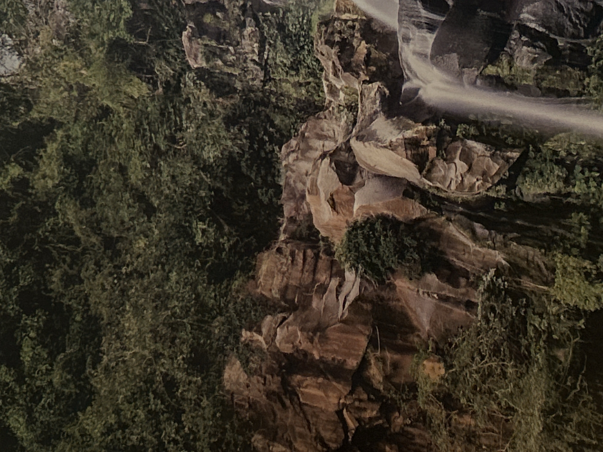The image shows a landscape with a waterfall cascading down a rocky cliff. Lush greenery covers the left side of the scene, contrasting with the brown and gray tones of the rock formations. The waterfall appears to be a thin stream of water. It is not possible to identify any specific location in Plovdiv, Bulgaria from this image. There are no visible people, subjects, or actions. The environment is rocky and vegetated with a waterfall feature.