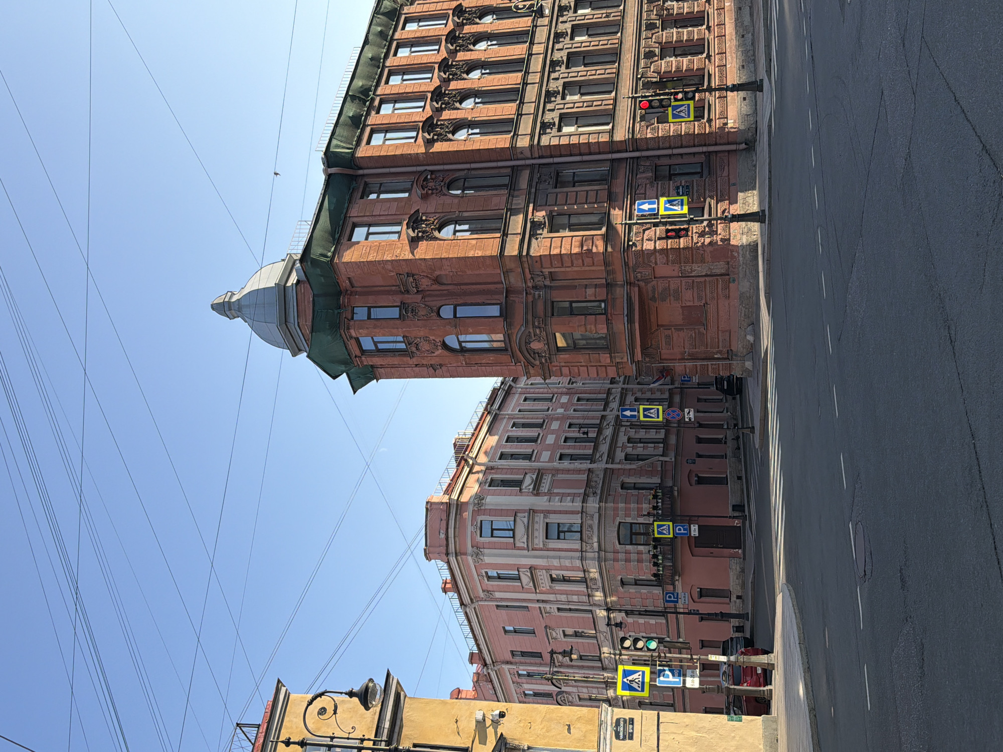 The image depicts a street intersection in Saint Petersburg, Russia, characterized by classic European architecture. Two prominent buildings frame the intersection: a taller, reddish-brown brick building with ornate detailing and a domed turret on the right, and a lighter pink, multi-story building with detailed cornices on the left. Both buildings appear to be residential or commercial, with numerous windows.
The foreground shows a paved street with pedestrian crossings marked by white stripes. Several traffic lights and street signs are visible. Blue pedestrian crossing signs with a white figure are prominent, along with traffic signals showing green and red lights. Yellow signs with the letter "P" indicate parking areas.
A red car is parked on the left side of the street, and a darker vehicle is partially visible behind it. The sky is clear and blue, with a network of overhead wires crisscrossing it. The scene appears to be captured during daylight, with the sun casting shadows. The overall impression is of a well-maintained urban environment with historical architectural elements.