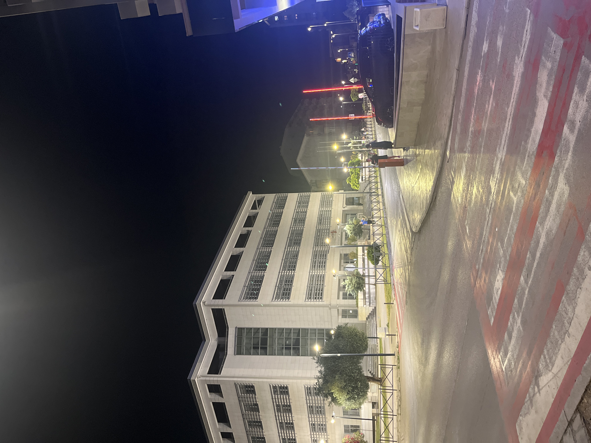 The image depicts a nighttime street scene in Budva, Montenegro. A modern, multi-story building with a light-colored facade and numerous windows occupies the left side of the frame. Streetlights illuminate the area, casting a warm glow on the wet pavement. A pedestrian crossing with red and white stripes is visible in the foreground. Several trees are planted along the sidewalk. In the middle ground, two vertical red lights are prominent, and a dark-colored car is parked. A few individuals are present on the sidewalk, some standing and others walking. The sky is dark and clear, indicating it is nighttime. The overall atmosphere is urban and somewhat tranquil, with the artificial lighting creating a reflective quality on the wet surfaces.
