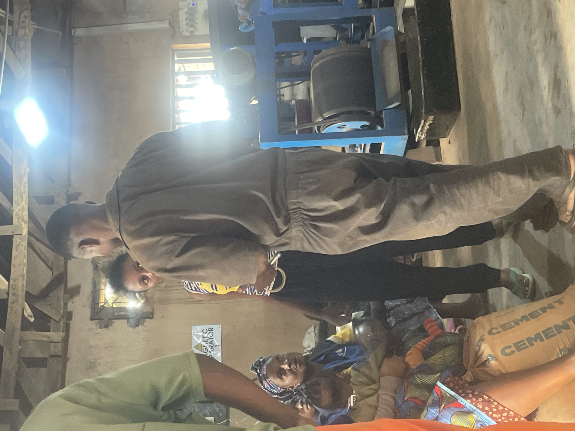 In an indoor setting resembling a workshop or industrial space in Gura Top, Nigeria, a group of people are gathered. A man wearing a brown uniform is speaking with a woman who is looking towards him. In the foreground, a woman is holding a baby, and a bag labeled "CEMENT" is visible. A blue industrial machine with a large roller is present in the background. The environment appears somewhat rudimentary, with exposed wooden beams in the ceiling and concrete walls. Natural light, supplemented by a bright overhead lamp, illuminates the scene. The overall impression is one of a community or work gathering in a functional, but not necessarily modern, setting.
