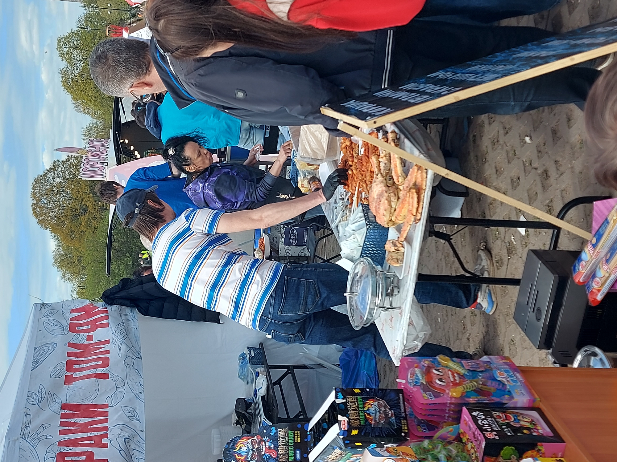 Here is a detailed description of the image:
The image depicts a vendor selling seafood, specifically what appears to be cooked crab and shrimp, at an outdoor market or event. A person wearing a striped shirt and a cap is tending to the seafood display. Another person with dark hair is also working at the stall. Customers appear to be browsing the seafood on display.
The background shows a tent or stall with the words "РАКИ ТОМ-ЯМ" written on it. There are various toys and merchandise displayed on a table below the seafood.
The environment seems to be an outdoor marketplace with several people in the background.
It is not possible to tell if the location is in the city of Saint Petersburg, Russia from the information provided.