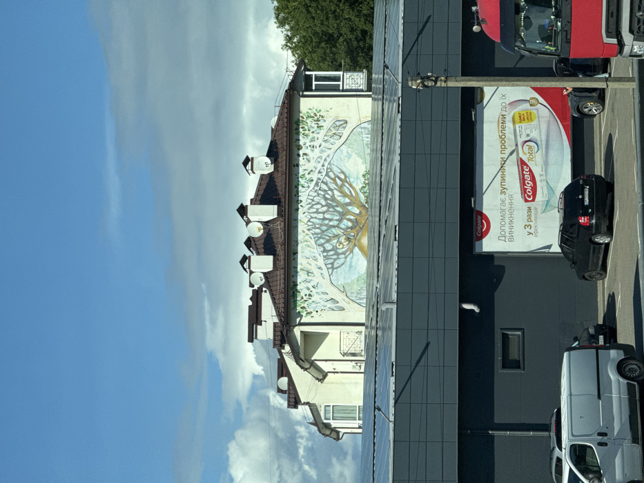 The image depicts an urban scene on Peremohy Street, Ukraine, under a partially cloudy blue sky during daylight. In the foreground, a modern, dark gray building features a flat roof covered with numerous solar panels. On its facade, a large billboard displays an advertisement for Colgate Total toothpaste in Ukrainian. A parking lot is visible in front of this building, occupied by a white cargo van, a black hatchback, another dark vehicle, and the partial front of a red semi-truck on the far right. A concrete utility pole stands in the right midground. Behind the dark gray building, a lighter-colored, multi-story building with a brown-tiled pitched roof is prominent. Its main facade features a large mural of a stylized tree with an owl perched within its branches, against a light blue and green backdrop. The roof of this background building is equipped with multiple brown chimneys and several white satellite dishes. Green trees are visible on the far right, behind the background building. No people are discernible in the scene.