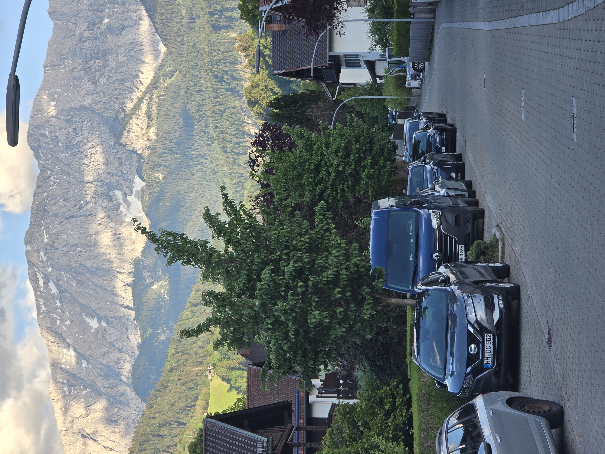 This image depicts a residential street in Garmisch-Partenkirchen, Germany, characterized by its Alpine setting. The scene is dominated by the majestic Wetterstein Mountains in the background, specifically the Zugspitze massif, with rugged grey peaks and visible snow patches, partially illuminated by sunlight under a sky with scattered white and blue clouds. Dense coniferous forests cover the lower mountain slopes, transitioning into lighter green hillsides with distant structures.
In the midground, traditional Bavarian-style houses with dark brown pitched roofs, white walls, and prominent balconies with dark green shutters or wooden details line the street. A particularly large, leafy green deciduous tree stands centrally on the left, partially obscuring houses and vehicles. Lush green hedges and various trees frame the street.
The foreground features a paved residential street made of interlocking grey pavers, with a white painted line along the right edge. A row of vehicles is parked on the left side of the street, facing away from the viewer. From closest to farthest: a silver station wagon (partially visible), a dark grey Nissan Qashqai SUV with a German license plate "HH DC 502" (Hamburg), a dark blue Renault Trafic van with a license plate "NH 1266", and several other dark cars. Modern, dark grey streetlights are visible on poles along the street. No people are visible in the scene.