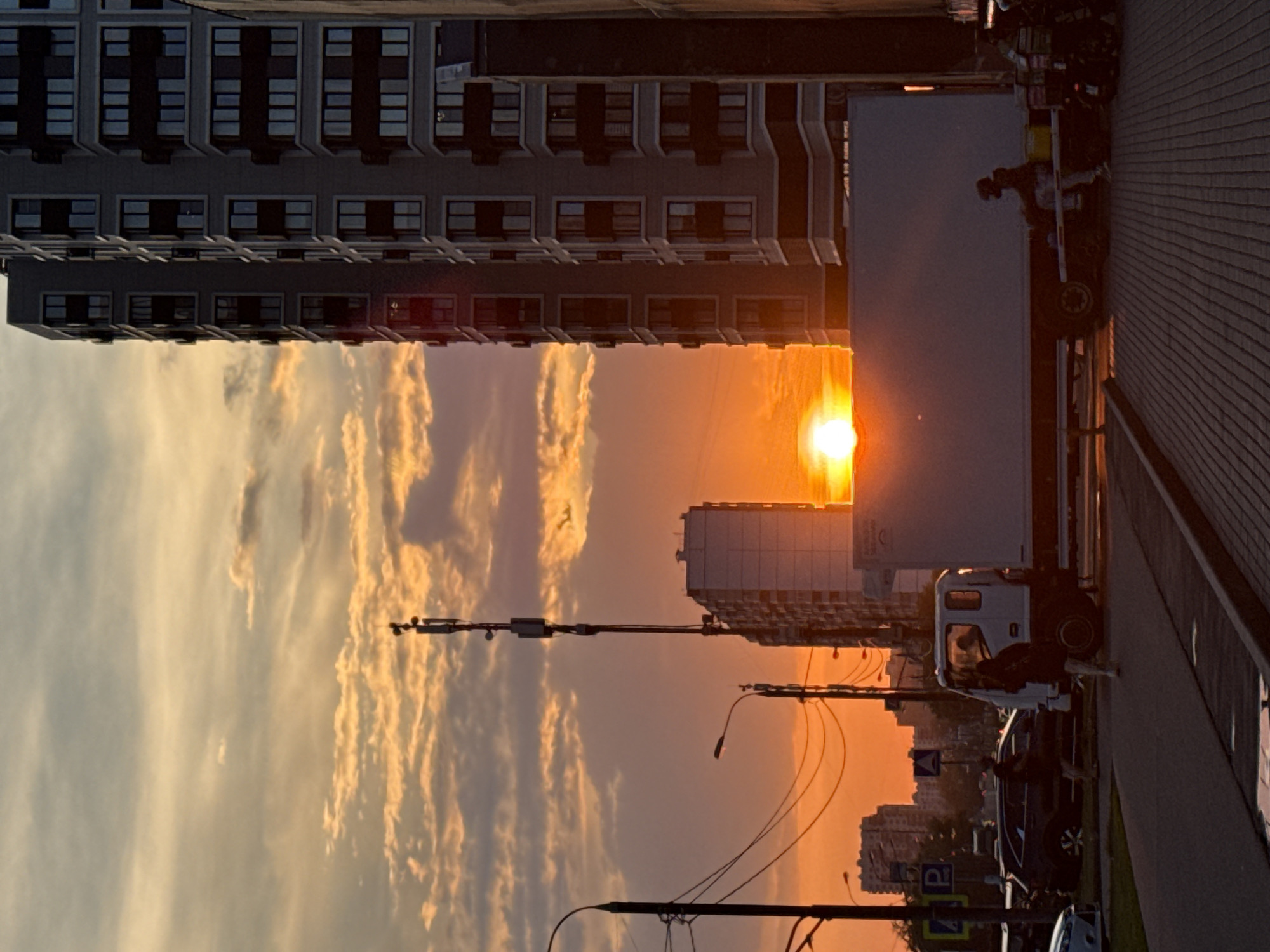 The image captures an urban sunset scene in Moscow, Russia. The sky is ablaze with orange and yellow hues, with scattered clouds creating a dramatic backdrop. In the foreground, a tall, modern residential building with numerous windows dominates the right side of the frame. To the left of this building, a telecommunications tower stands tall, its antennas silhouetted against the sky.
Below the tower, a white box truck is parked, its side reflecting the warm light of the setting sun. Two individuals are visible near the truck: one person is standing on the sidewalk, and another is seated on the truck's chassis. Further to the left, a dark-colored SUV is parked, and two more people are walking on the sidewalk. Traffic lights and street lamps are also present, indicating a busy urban environment. The ground in the foreground is paved with textured material, possibly a sidewalk or pedestrian area. The overall impression is a snapshot of city life at dusk, with the dramatic natural light softening the urban landscape.