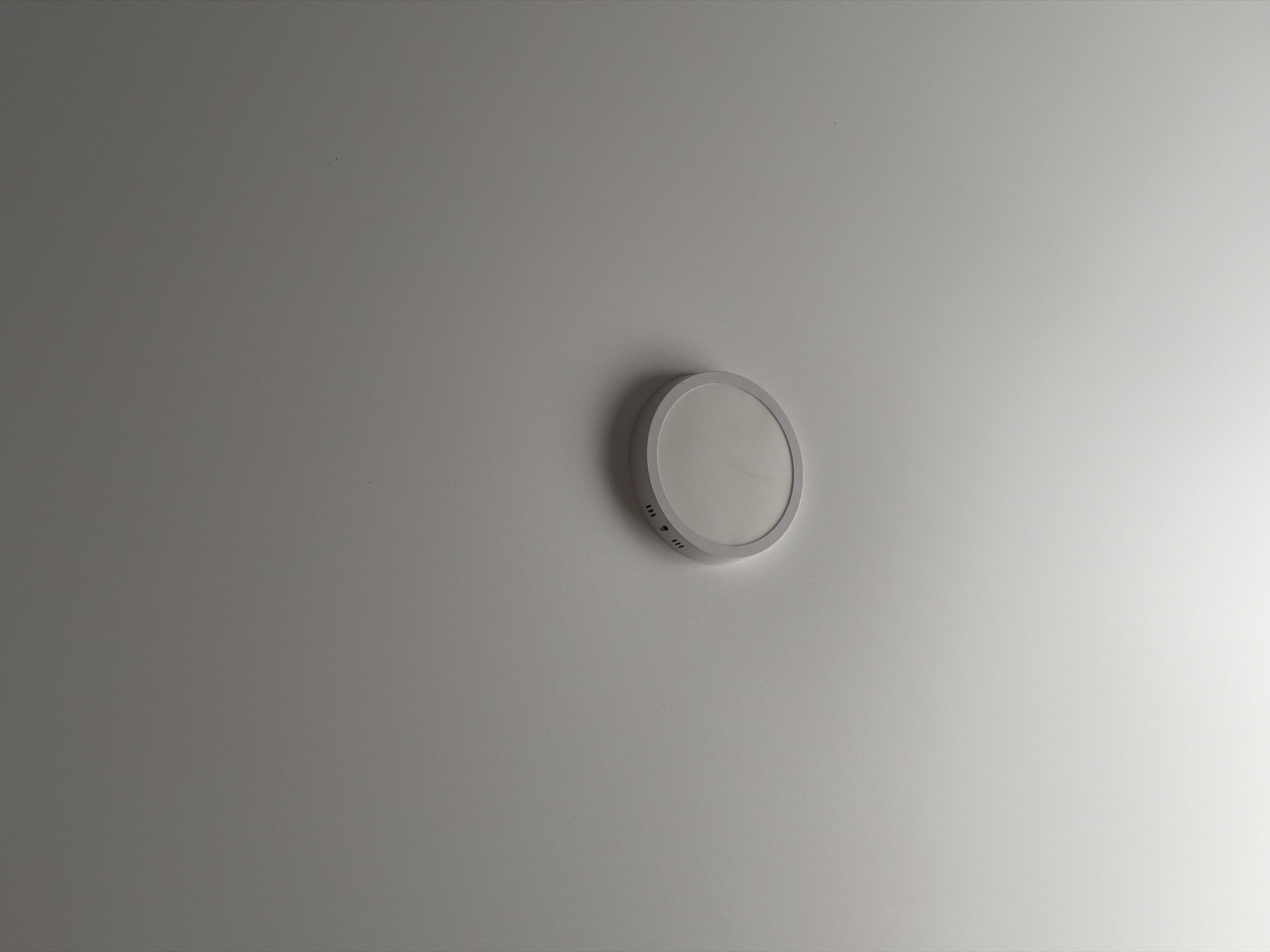 The image shows a round, white ceiling light fixture installed on a white ceiling. The fixture has a slightly raised, metallic rim and a frosted diffuser. Small, discreet ventilation holes are visible around the edge of the fixture. The lighting is soft and even, suggesting the light is on. The environment appears to be an indoor space, likely a room or hallway, with no other objects or people visible in the frame. The image does not contain any information that would allow for its localization in the city of Strășeni, Moldova.