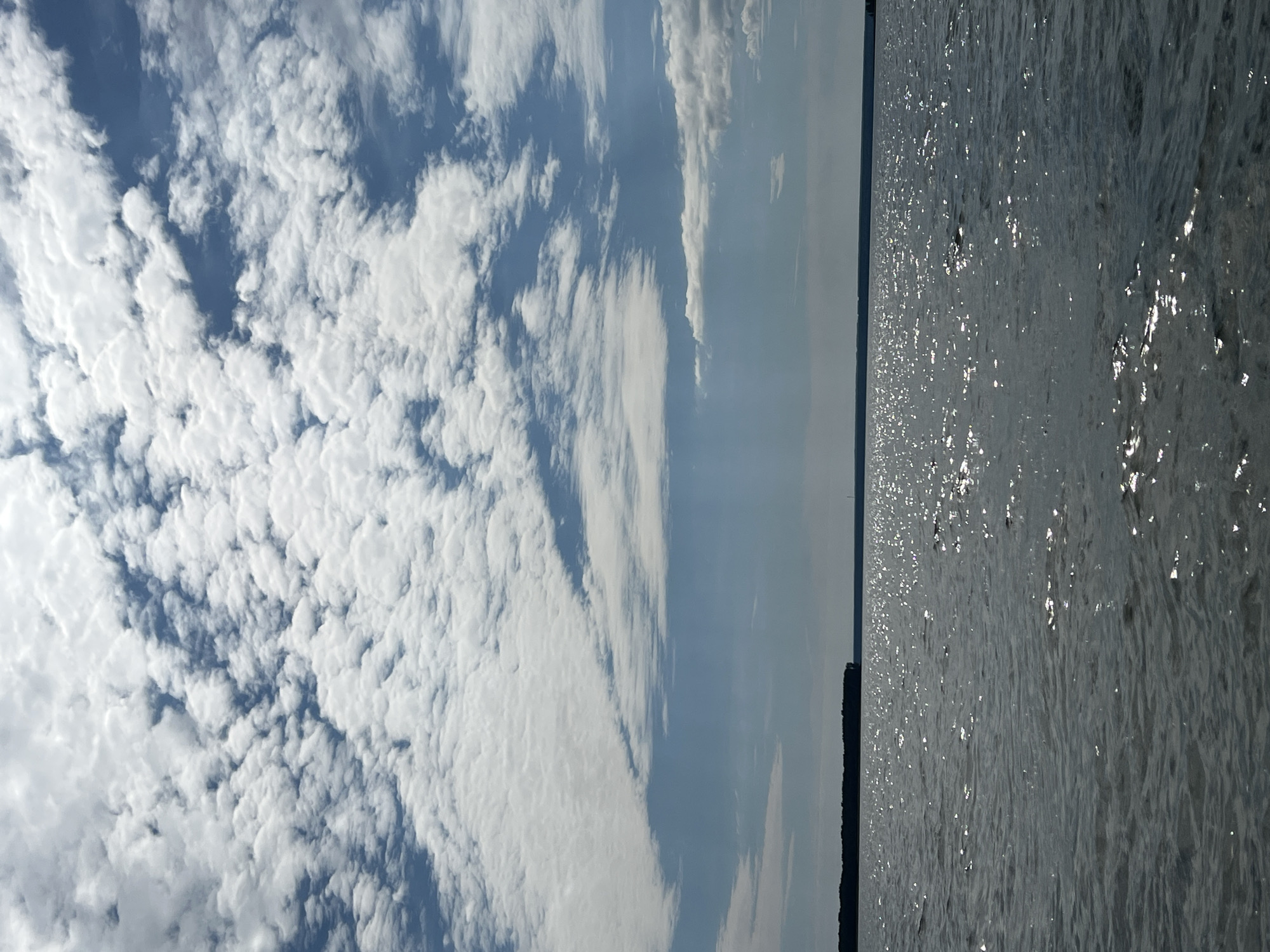 The image depicts a wide expanse of shimmering water under a partly cloudy sky. In the distance, a dark, tree-lined horizon is visible, suggesting a natural landscape. The water's surface is textured with ripples and reflects the sunlight, creating a dappled effect. The clouds are a mix of white and gray, interspersed with patches of blue sky. There are no people or specific man-made structures identifiable in the scene. The location is not specified within the city of Novopichugovo, Russia.