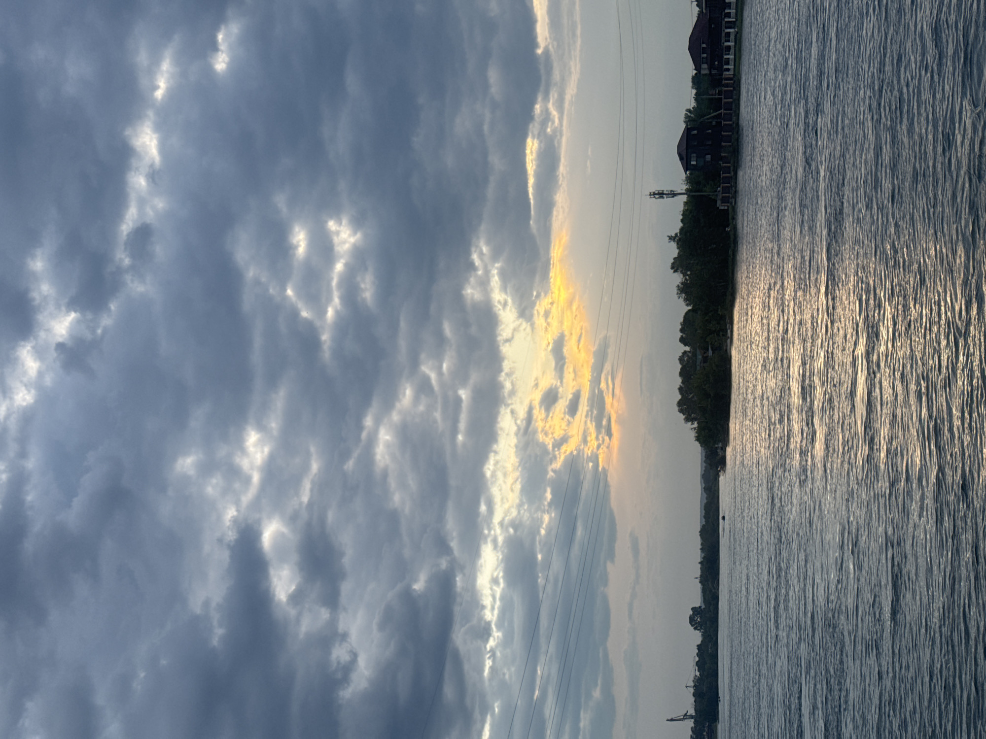 The image depicts a body of water, likely a river or lake, under a dramatic, overcast sky. Dark, heavy clouds dominate the upper portion of the frame, with a break in the clouds allowing a sliver of bright, yellowish sunlight to shine through. The water's surface is rippled, reflecting the subdued light. Along the far bank, a line of trees and some buildings are visible. Among the structures, there appear to be houses with dark roofs and a communication tower. Power lines stretch across the sky in the middle ground. In the distance, on the left side of the frame, a large industrial crane is partially visible. The overall impression is one of a slightly somber, natural landscape with hints of human habitation. This scene is consistent with views along the Angara River in Irkutsk, Russia, given its geographical features and industrial elements. There are no people or discernible animals visible in the image.