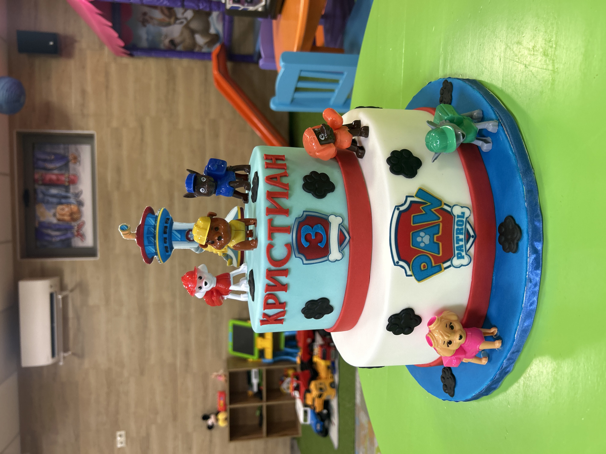 The image shows a two-tiered Paw Patrol themed birthday cake, decorated with figurines of the Paw Patrol characters and the name "КРИСТИАН" (Kristian) and the number "3". The cake has a blue and white color scheme with red accents. The cake is placed on a green table. The background is a playroom setting with toys, furniture, and wall decorations. The location is not explicitly evident from the image. The image is taken indoors. The city of Plovdiv, Bulgaria is not identified in the image.