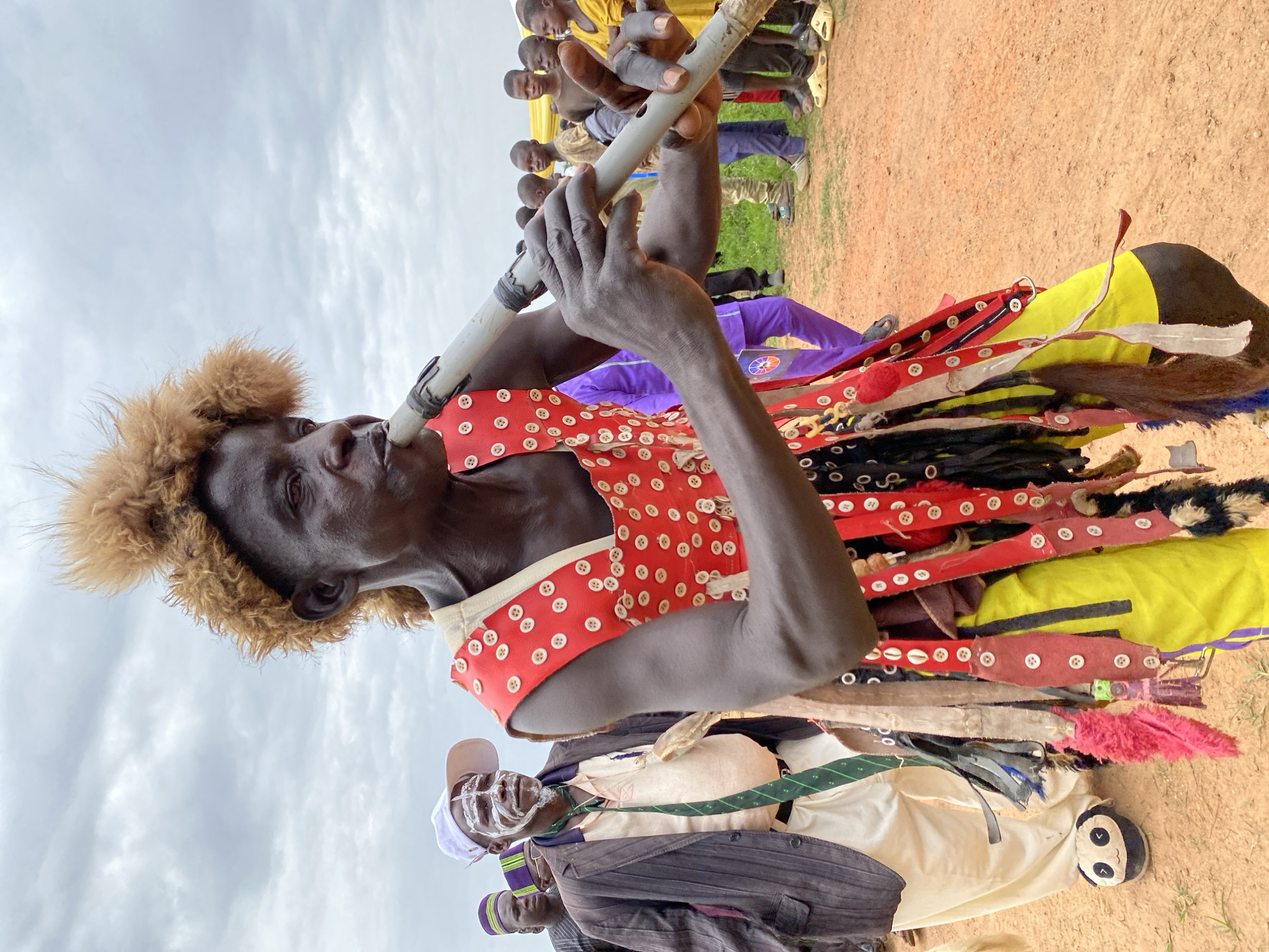 The image depicts an individual playing a long, cylindrical flute. The person is dark-skinned, has short curly hair, and is wearing a red outfit adorned with white buttons and colorful stripes. The individual appears to be outdoors, on a dirt ground, with other people standing behind. The setting includes a cloudy sky. The location is Kafanchan A, Nigeria.