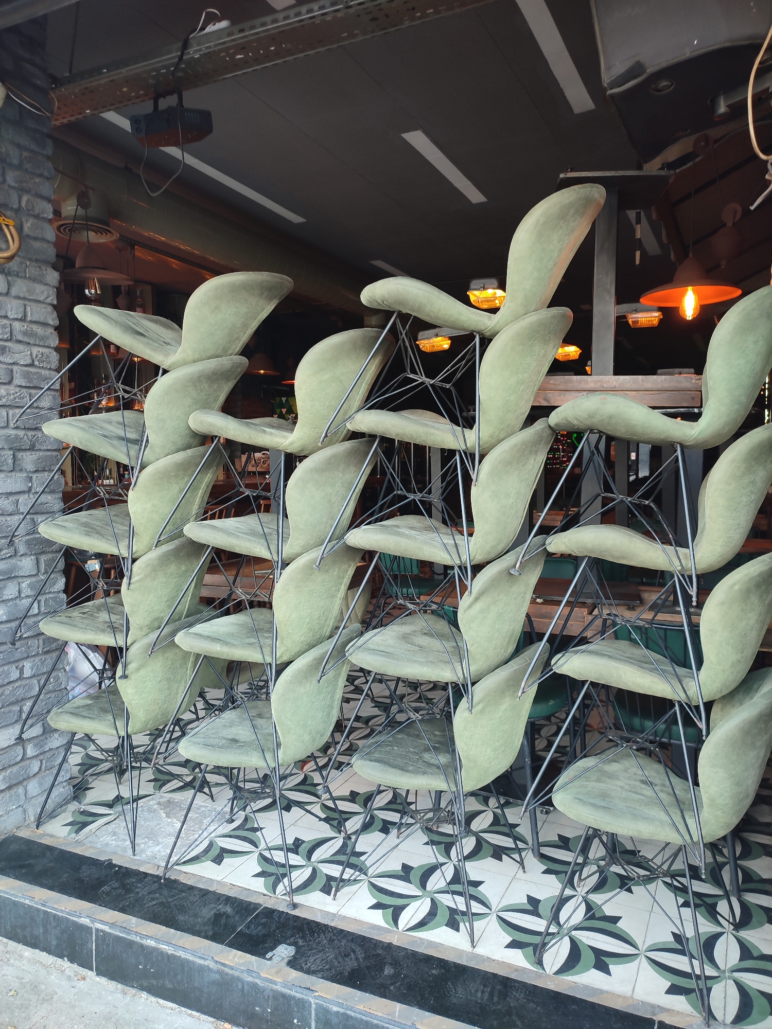 The image shows a stack of light green chairs with black metal frames, piled high on a patterned tile floor. The chairs appear to be outdoors, possibly at the entrance to a cafe or restaurant, as suggested by the exposed ceiling beams, lighting fixtures, and brick wall. The overall impression is one of a commercial establishment preparing for or closing down for the day. The scene is likely located in Kadıköy, a vibrant district on the Asian side of Istanbul, Turkey, known for its lively cafes and street life.