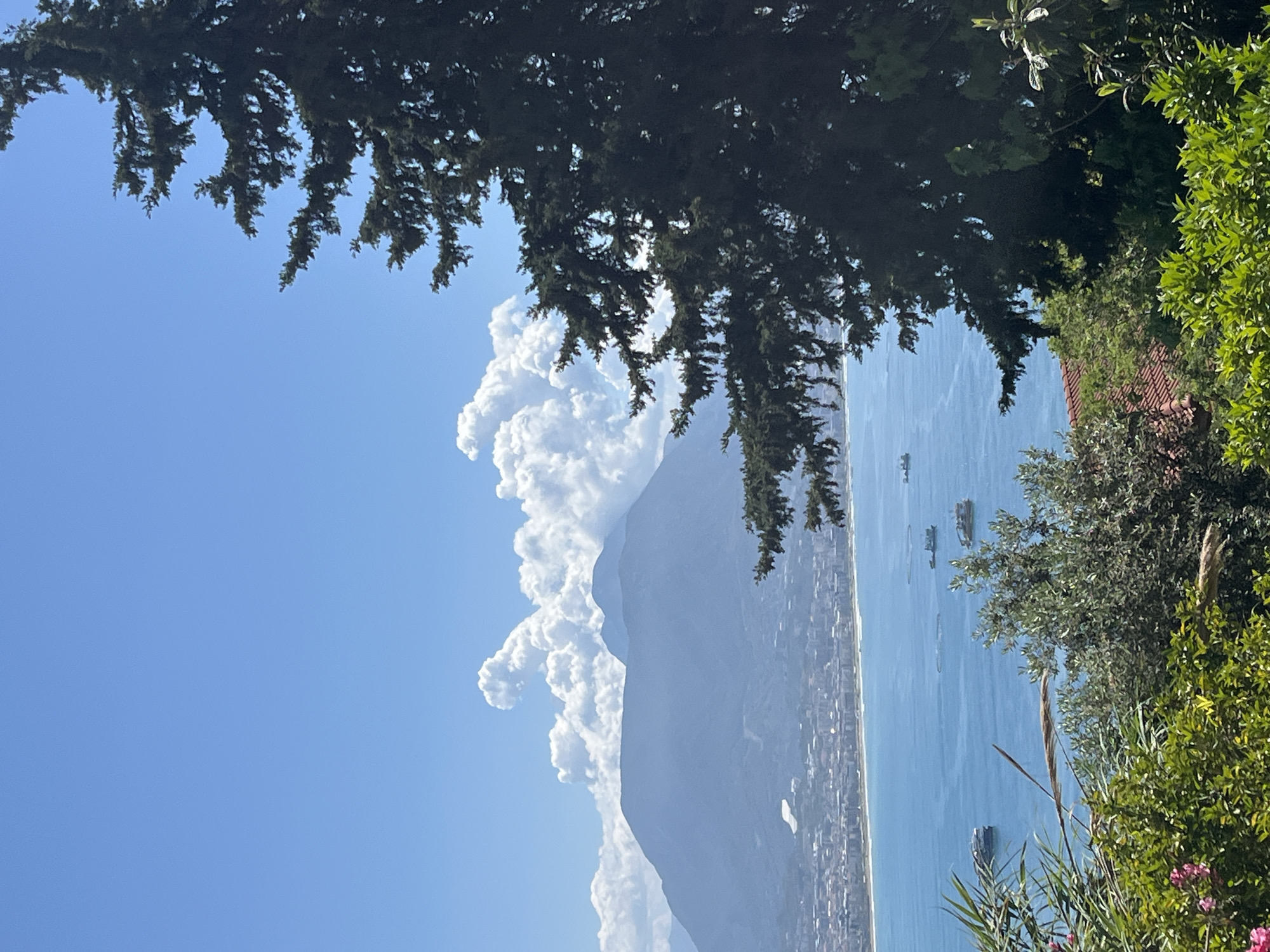 The image captures a view from a vantage point overlooking Alanya, Turkey, or a nearby coastal area. Dominating the left side is a mountain shrouded in cumulus clouds under a blue sky. In the middle ground, a city stretches along the coastline, bordering the sea. The sea itself appears calm with several boats visible. In the foreground, branches of a tree and other lush greenery frame the top and right side of the shot. The image is taken on a clear, sunny day.