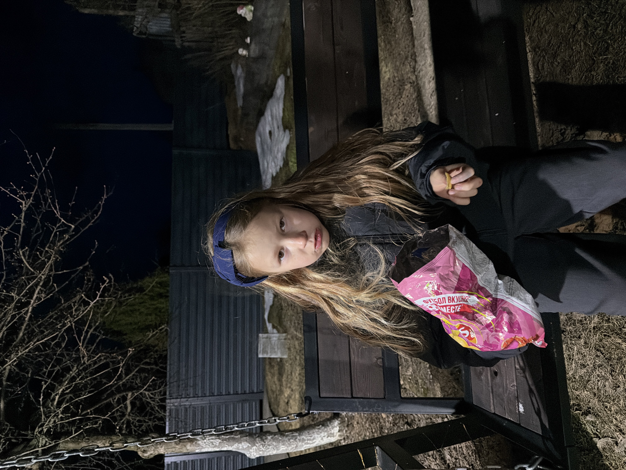 A young girl with long blonde hair sits outside in Dubovaya, Russia, dressed in dark clothing and a blue headband. She is holding a chip in one hand and a crumpled pink chip bag in the other. A bare tree and a chain are visible in the background. The scene appears to be at night.