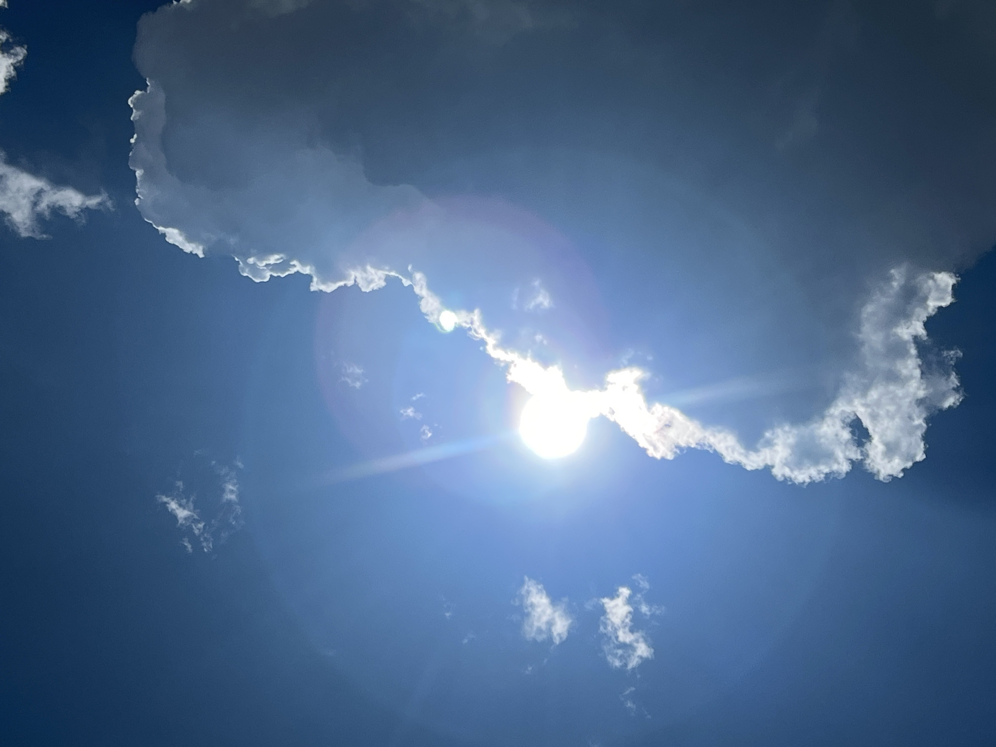 This image is a close-up, low-angle shot of a bright sun partially obscured by a large, dark cloud formation. The sun is a brilliant, diffused sphere emitting strong rays that create lens flare and a visible halo effect. The cloud to the right is dense and textured, with its edges illuminated by the sunlight, creating a contrast between light and shadow. Smaller, wispy white clouds are scattered across the deep blue sky. The scene appears to be outdoors, with no human subjects or specific objects visible other than the clouds and the sun. The location of Mochishche, Russia is not depicted in the image.
