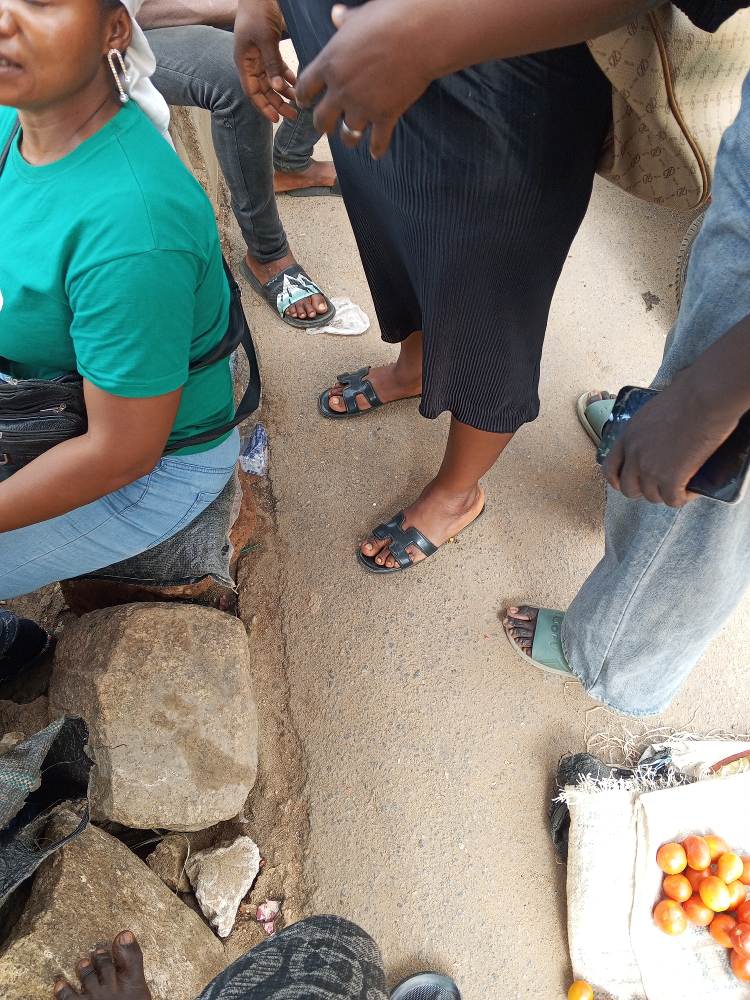 The scene is an outdoor street setting in what appears to be the city of Gwong, Nigeria.
There are multiple individuals present: a woman is seated on a stone structure, wearing a green t-shirt, light blue jeans, and an earring. Other people surround her, including a person with black slacks and another with denim jeans. Each are wearing flat sandals.
Objects include a black purse/bag on the seated woman, several rocks that are being used as the seat, a plastic bottle, and what looks like a display of tomatoes on a piece of white fabric.
The surface is concrete or paved.