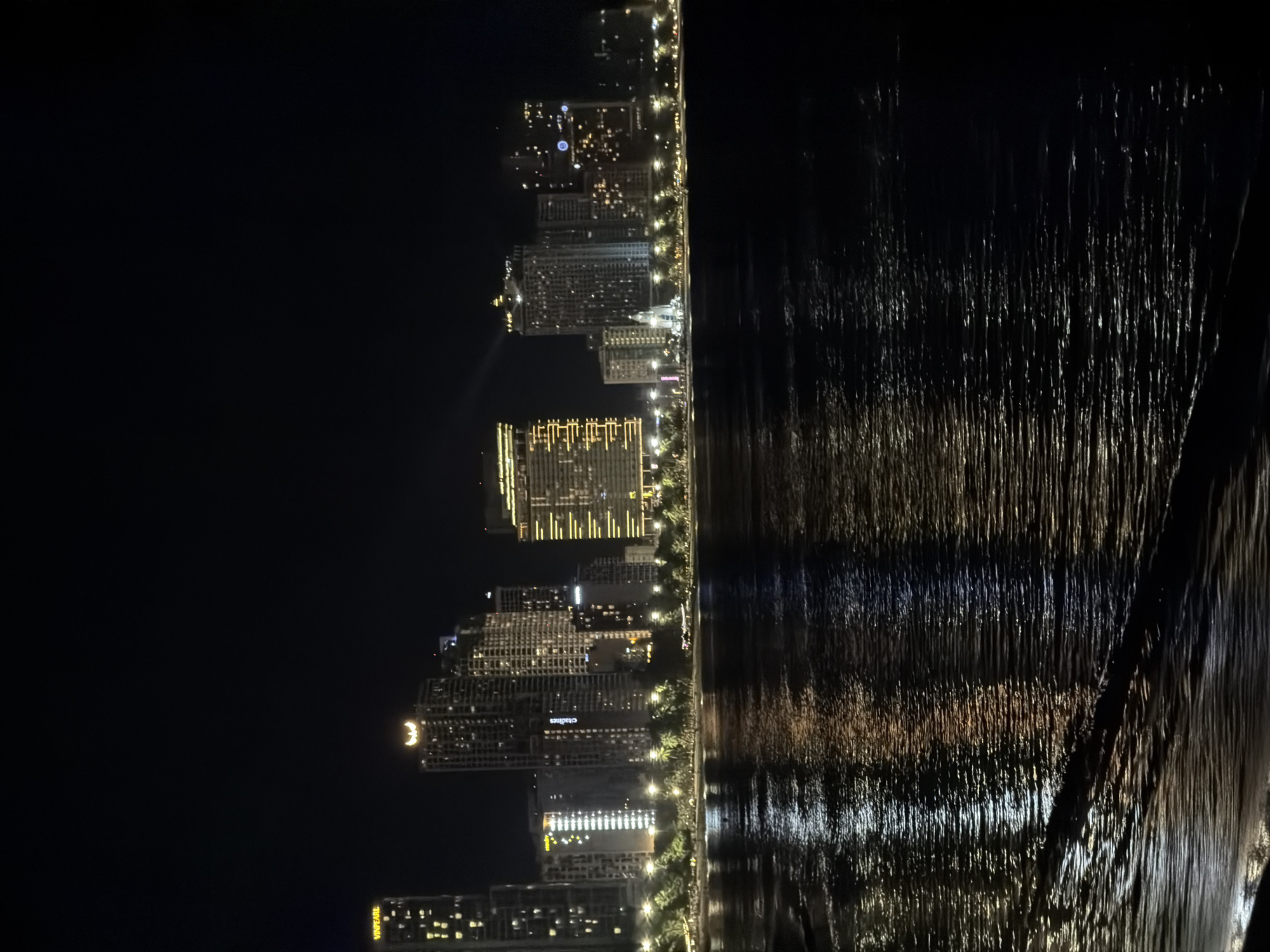 This is a nighttime photograph of the Nha Trang waterfront in Nha Trang Ward, Vietnam. The scene is dominated by a dark, choppy sea in the foreground, reflecting the lights of the city skyline in the background. The skyline features a collection of modern, illuminated high-rise buildings, including hotels and apartments. Some buildings display distinct architectural features and lit signage, such as "VINPEARL" on a tower to the far left, and another building with a crescent moon-like emblem on top. Palm trees are visible lining the waterfront promenade, interspersed with streetlights. The overall impression is that of a vibrant, developed coastal city at night. There are no people or animals visible in the image.