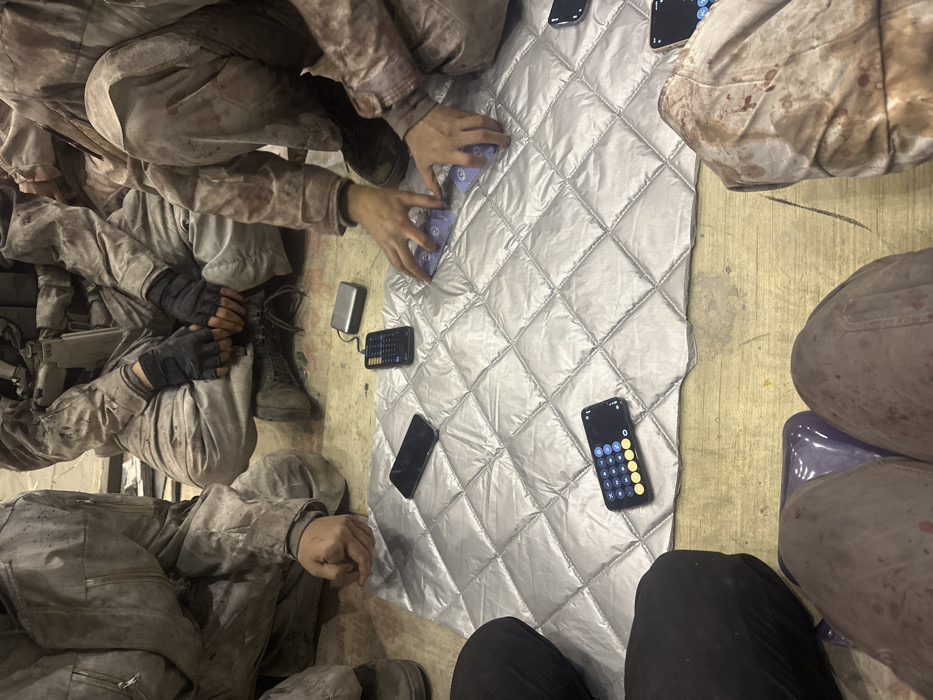 The scene, located in Huangdao District, China, captures five individuals from a high-angle perspective, gathered indoors on a light-colored floor around a silvery, quilted mat. Three individuals are dressed in camouflage uniforms heavily marked with reddish-brown liquid, consistent with simulated blood. One individual on the upper right wears dark tactical gloves, and a visible "UEB" patch is affixed to their vest or equipment. Another individual on the lower left wears plain dark clothing. A fifth individual's lower legs are visible at the bottom of the frame, resting on what appears to be purple footwear or a cushion.
On the quilted mat, four smartphones are present: one is face down, two are actively displaying calculator interfaces, and a third is connected via a cable to a small black device resembling a portable keyboard. A silver portable electronic device, likely a hard drive or power bank, rests near the connected smartphone. The individual on the right is actively manipulating a stack of blue-backed playing cards featuring a distinct eye-motif design. The floor is light-colored and exhibits wear and scuff marks. The background is undifferentiated, indicating an enclosed, utilitarian environment.