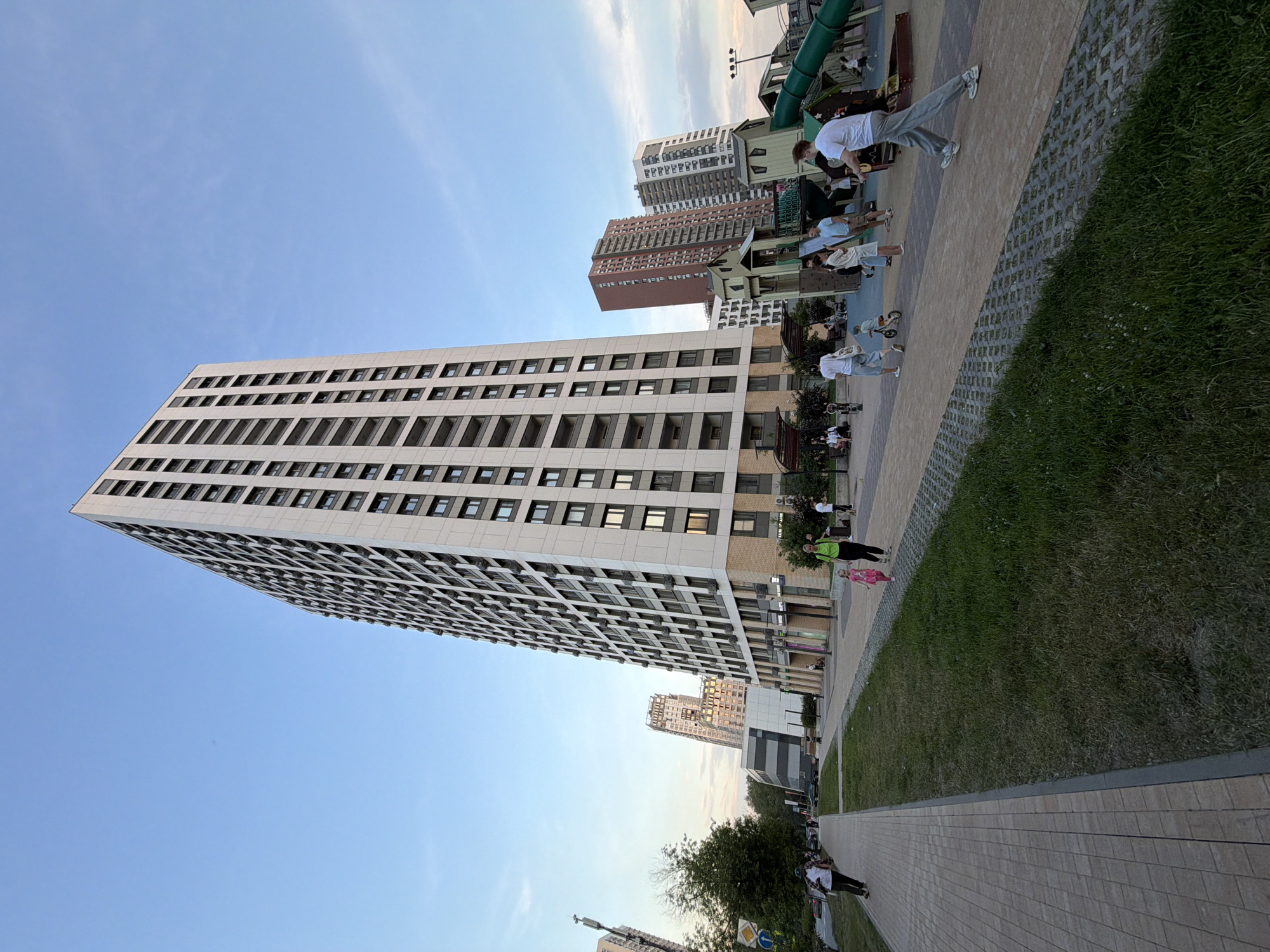 In a cityscape in Moscow, Russia, a towering, modern residential building dominates the frame, its white facade punctuated by numerous dark-framed windows and balconies stretching towards a clear blue sky. The building's base is a lighter brick color, and it appears to be part of a complex of contemporary high-rise apartments, with other buildings of varying heights and colors visible in the background.
In the foreground, a paved walkway is bordered by a strip of lush green grass. Several people are present in the scene. A woman in a bright green shirt and black pants walks alongside a young girl in a pink dress. Further down the path, a man on a skateboard moves away from the camera, while another man walks with a dog. To the right, a playground area is active with children and adults. A man in a white t-shirt and grey pants is captured mid-stride. Several children are playing on the playground equipment, which includes slides and climbing structures designed in a whimsical, house-like style. The scene appears to be set in the late afternoon or early evening, with the sky casting a soft light.