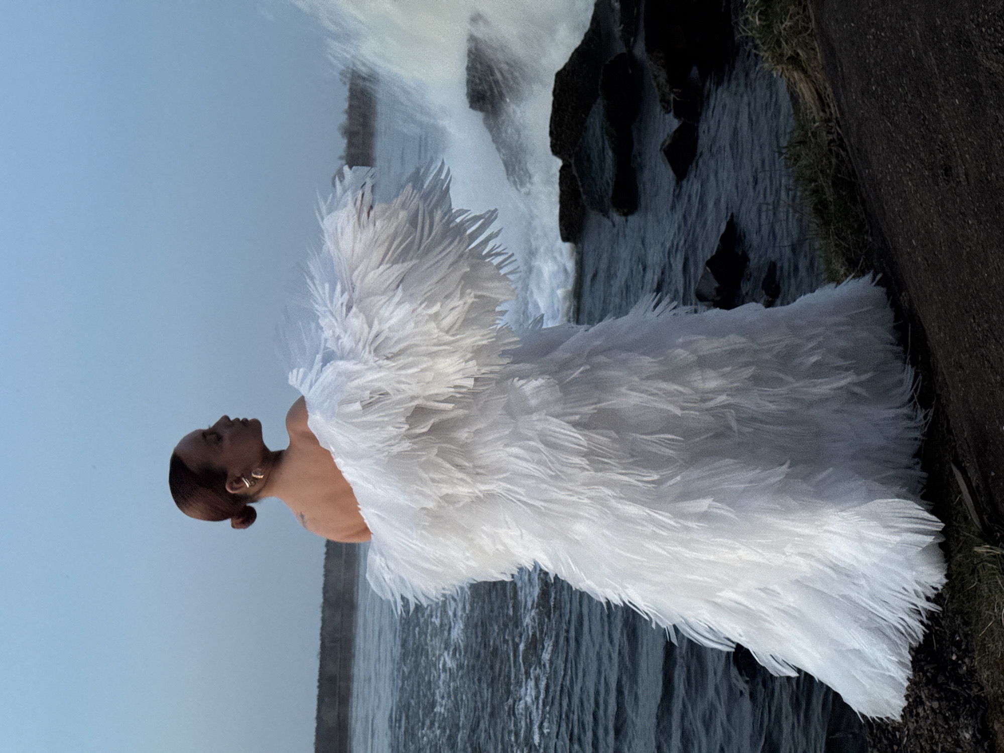 A woman wearing a white, feathery garment stands in front of what appears to be a waterfall and rocky shoreline. The woman has dark skin, her hair is styled in a bun, and she is wearing a ring in her ear. The background features a waterfall that rushes down into a dark body of water. The setting appears to be natural and possibly near a rocky bank or shore. It is not possible to confirm the location in Chaikovsky, Russia, without additional information.