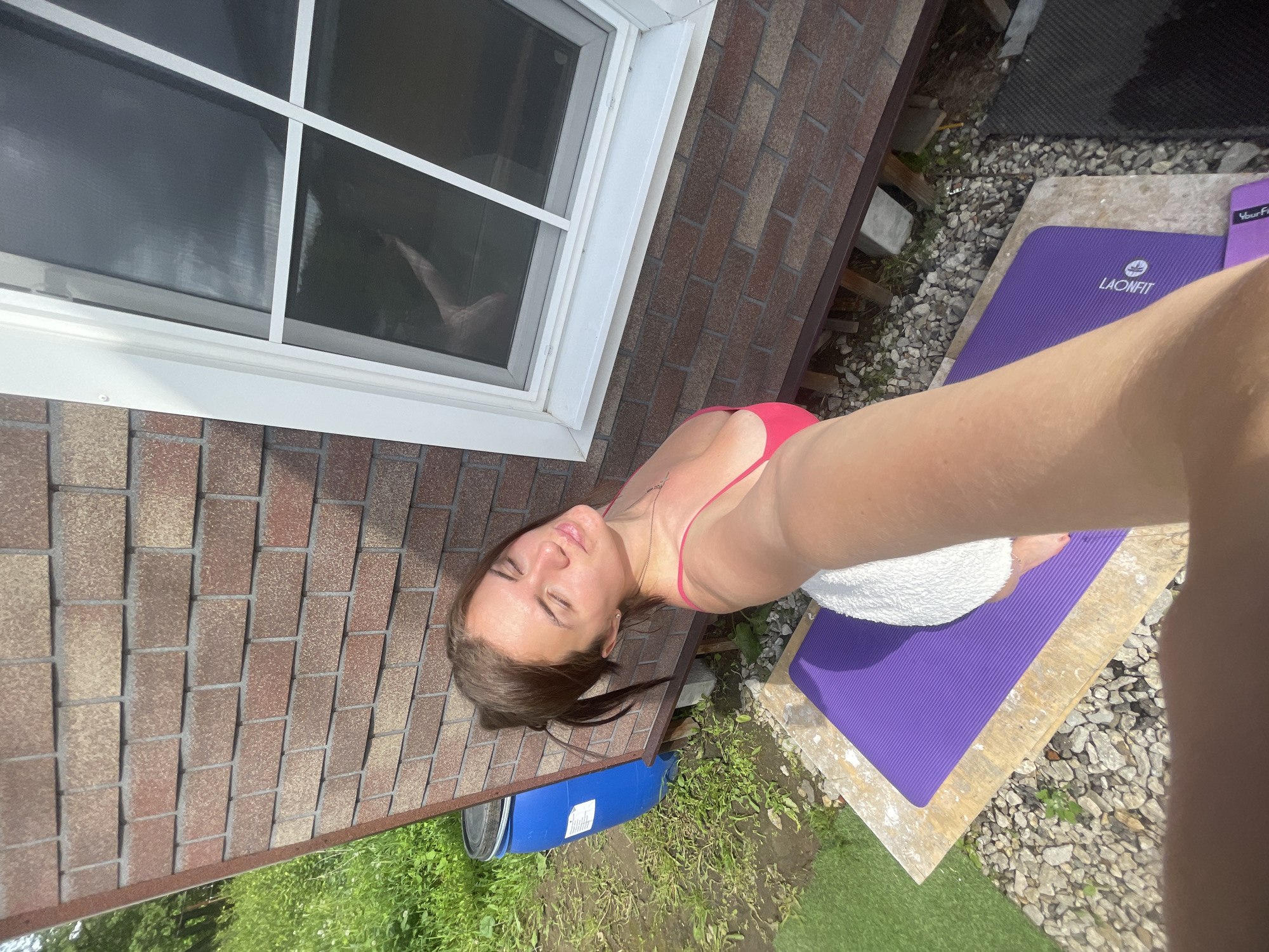 A woman in a red swimsuit is taking a selfie outdoors. She is positioned next to a house with brick siding and a window. A blue barrel is visible on the ground nearby. A purple yoga mat is laid out on a platform surrounded by rocks and grass. The location is likely in a residential or outdoor area, but specific details regarding the city or exact location in Мосметаллоконструкция Онт, Russia cannot be definitively determined from the image alone.