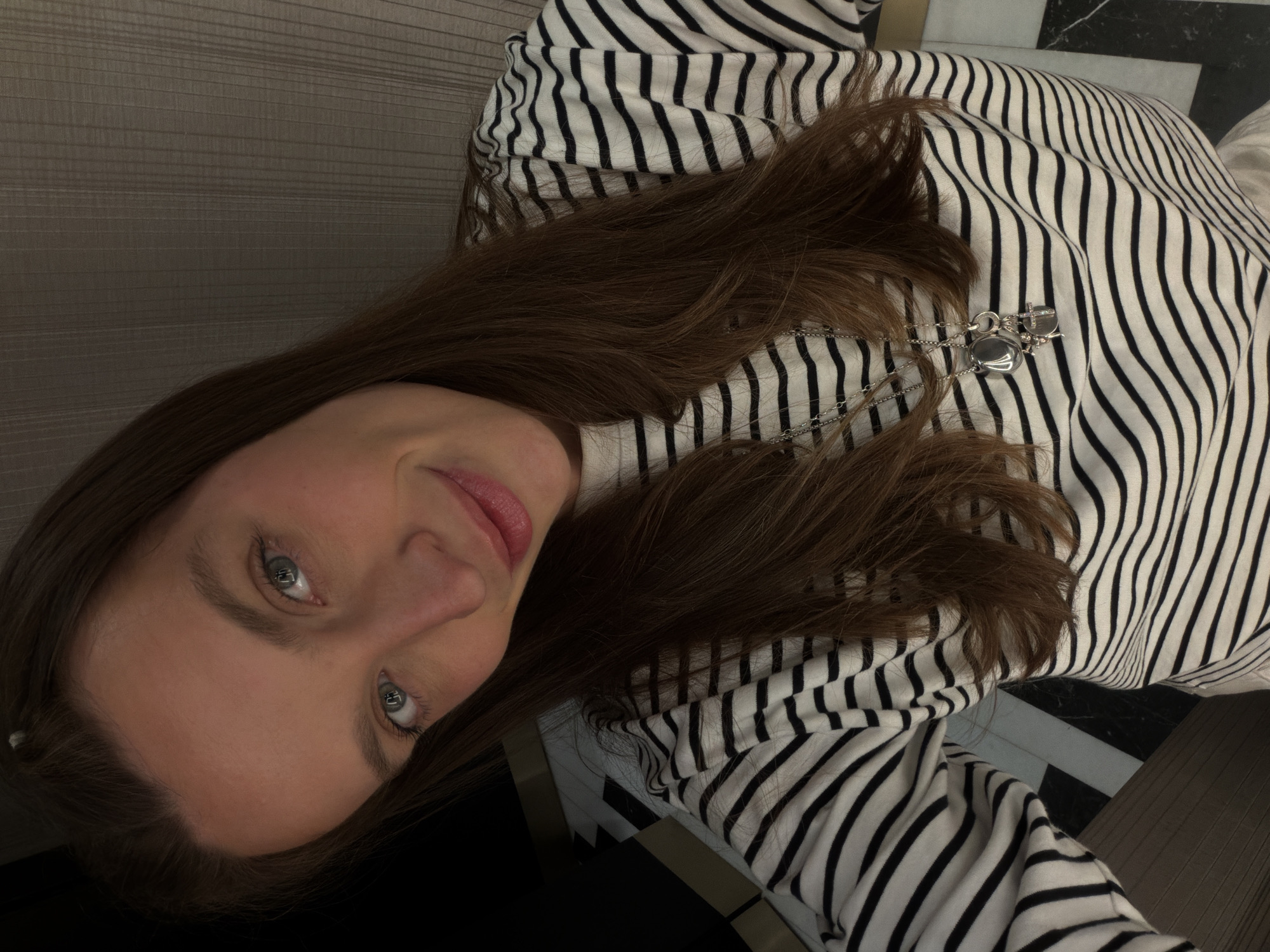 A woman with light skin, brown hair, and blue eyes poses in a close-up shot. She wears a black and white striped shirt and a silver necklace with several pendants. The background shows a wall, a floor, and possibly furniture. The floor appears to have a black and white tile pattern, suggesting an interior space. The image location cannot be determined from the given information.