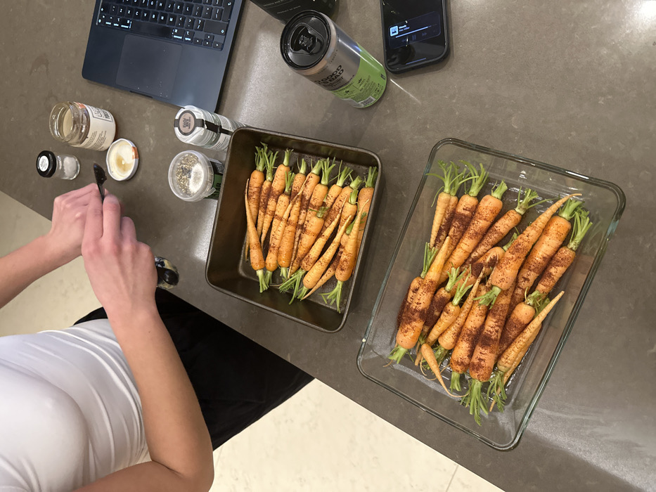 A woman in a white top prepares carrots on a gray countertop. Two baking dishes, one metal and one glass, contain rows of orange carrots with green tops, sprinkled with a reddish-brown spice. Nearby are jars of spices and other ingredients, along with a laptop, a green can, and a mobile phone. The scene suggests a kitchen or food preparation area.