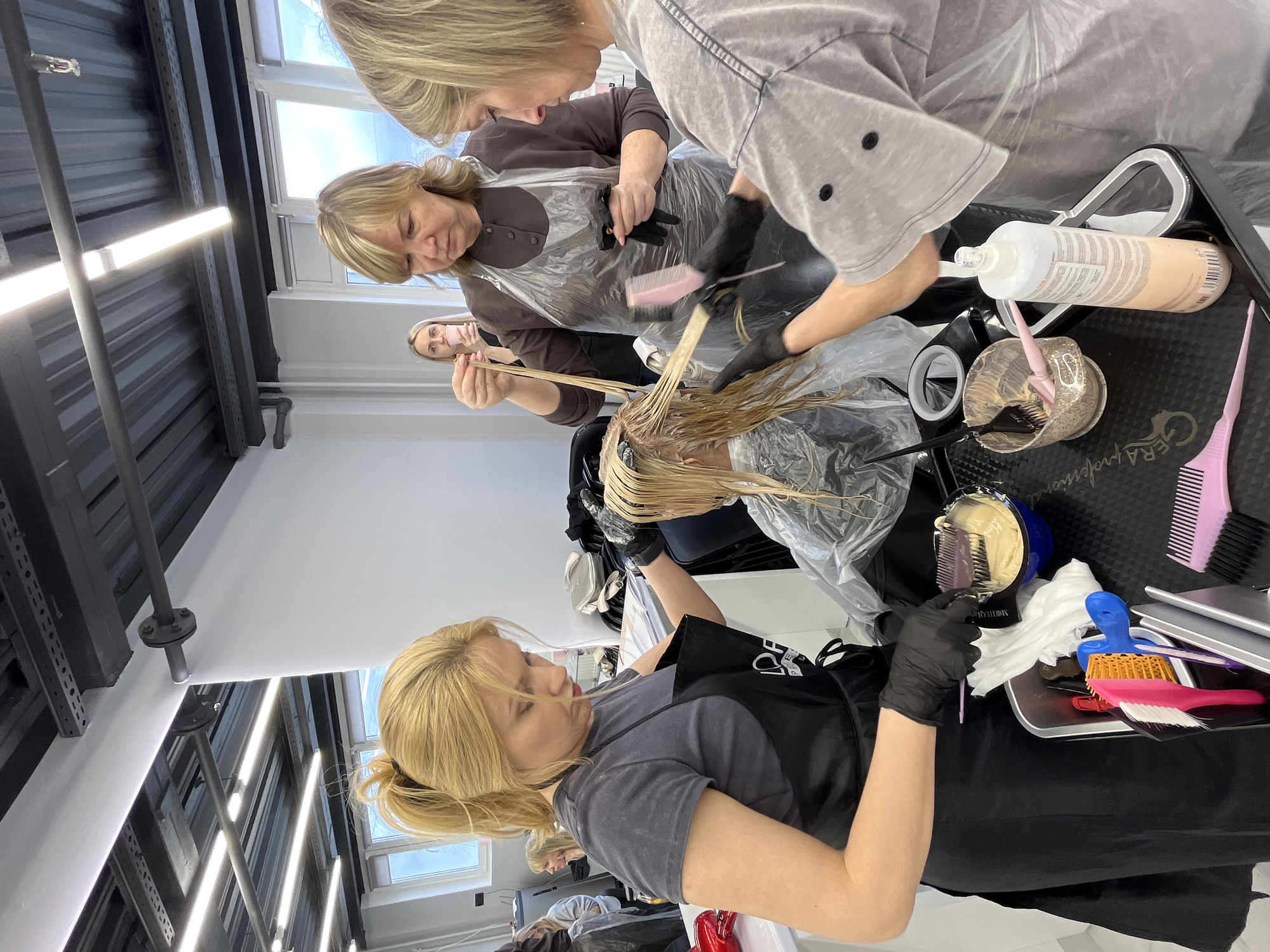 This media file captures a hair coloring or lightening session in a professional salon environment in Izhevsk, Russia, likely during the daytime due to ample natural light from large windows.
The scene features three women actively engaged in treating a client's blond hair. In the immediate foreground, a woman with blond hair in a ponytail, wearing a grey t-shirt, black apron, and black gloves, holds up a section of the client's hair. Behind her, a second woman, also blond, dressed in a brown cardigan and a clear plastic smock, meticulously applies hair product to the client's hair using a pink brush while wearing black gloves. A third blond woman, wearing a light-colored top and possibly a face mask, is partially visible in the background, observing or assisting. The client, whose back is towards the camera, is seated in a black salon chair, covered by a clear plastic cape, undergoing the hair treatment.
Various hair styling tools are organized on a black utility tray beside the stylists. These include several brushes (pink, white, orange/yellow), pink and purple combs, and mixing bowls containing hair product. One mixing bowl has text that appears to be "MONTE BELLU," while a white bottle of hair product is clearly labeled "PROFESSIONAL." The black tray itself bears the text "GERA Professional."
The salon's interior features an industrial-style ceiling with exposed black metal beams and ducts, complemented by bright fluorescent strip lighting. The walls are white, and large windows provide a view to the outside. The overall atmosphere suggests a focused and professional setting.