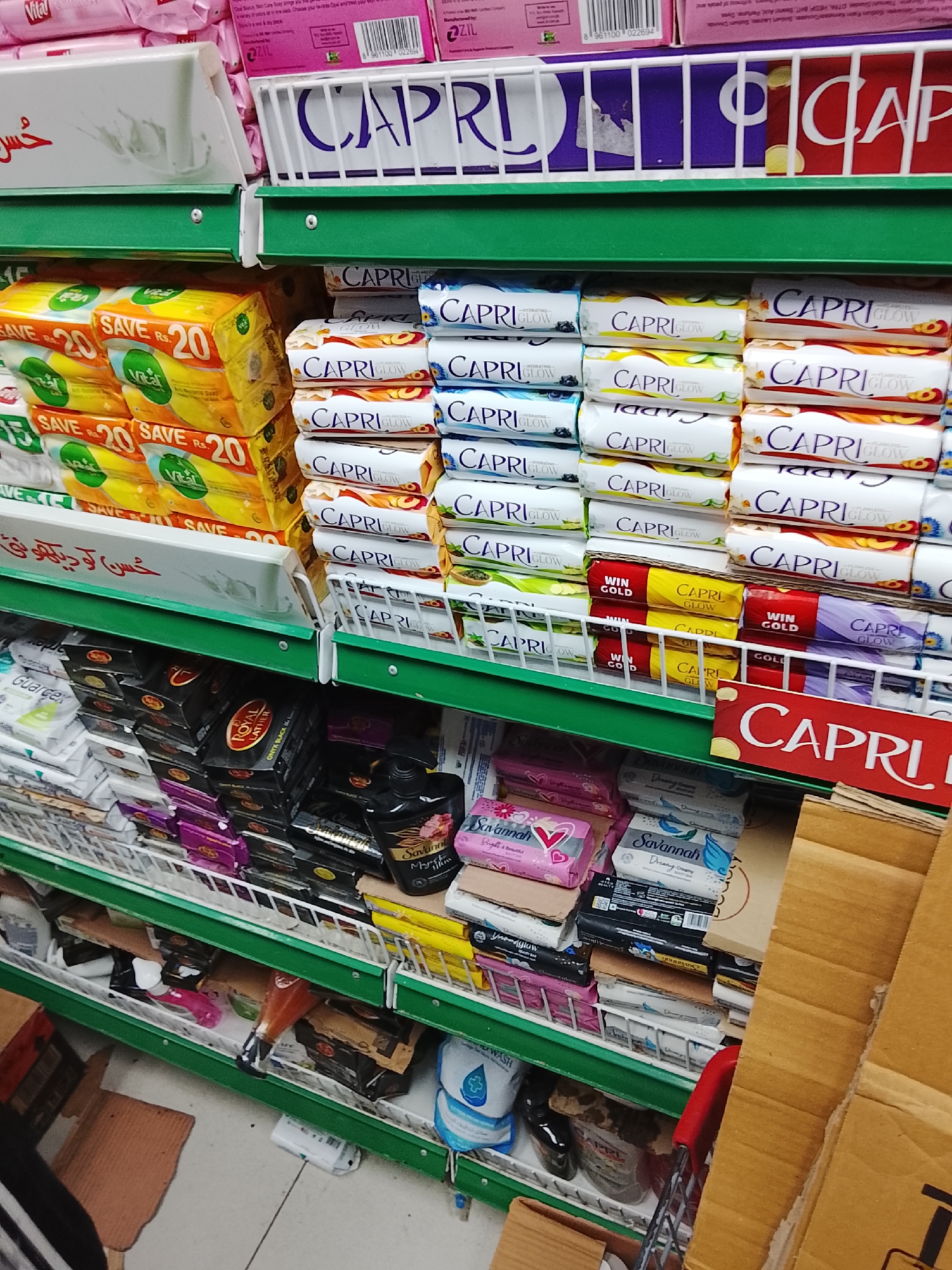 The image depicts a shelf in a store, likely located in the Gulgasht Colony in Pakistan, based on the Urdu script on some of the products. The shelves are stocked with various brands and types of soaps, including Capri, Vital, Savannah, Royal Lather, and Guarcex. The Capri soaps come in different varieties and packaging colors. A shopping cart is visible near the bottom right of the frame. There are also other household and personal care items on the shelves.