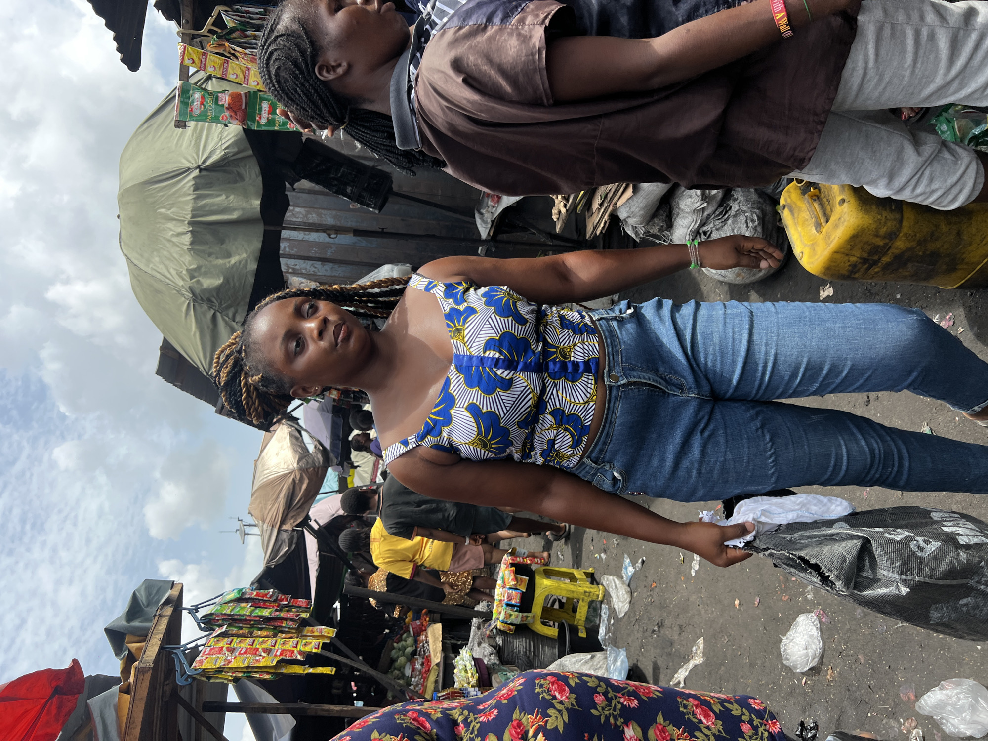 The image captures a street scene in Nkpor, Nigeria, with two women prominently featured in the foreground. The first woman is wearing a blue and white floral print tank top and blue jeans. Her hair is styled in braids. She is holding a black plastic bag with white fabric or items inside. The second woman, partially visible at the top, wears a brown button-down shirt, gray pants, and has her hair braided. She also wears a beaded bracelet.
The background reveals a bustling marketplace. Various vendors are present, selling goods from stalls or tables, some covered with umbrellas or tarps. Merchandise, including packets of seasoning or spices, are displayed. A yellow plastic stool is visible. The ground appears to be concrete or pavement and is littered with debris, including plastic bags. Other people are visible in the background, some in yellow shirts, further indicating a crowded, public space.