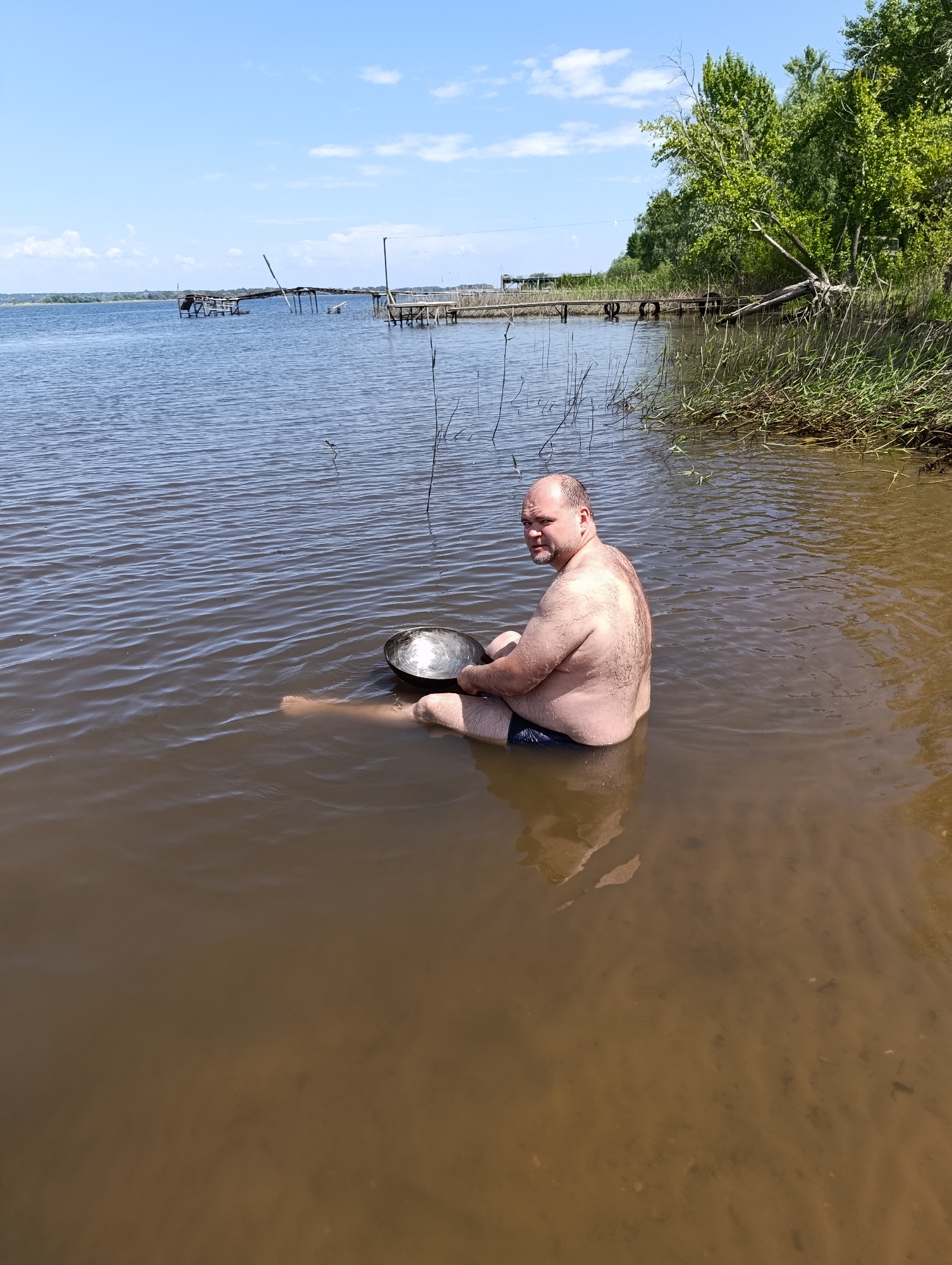 A shirtless, overweight, bald man sits in shallow, murky water, holding a metal pan. He is wearing dark swim trunks. The man is facing away from the camera, looking over his left shoulder. He is seated with his legs extended in front of him. The water is calm with ripples on the surface. In the background, there are wooden docks and structures, as well as trees and reeds. The sky is blue with scattered white clouds. The scene appears to be a natural body of water, possibly a lake or river, on a sunny day.