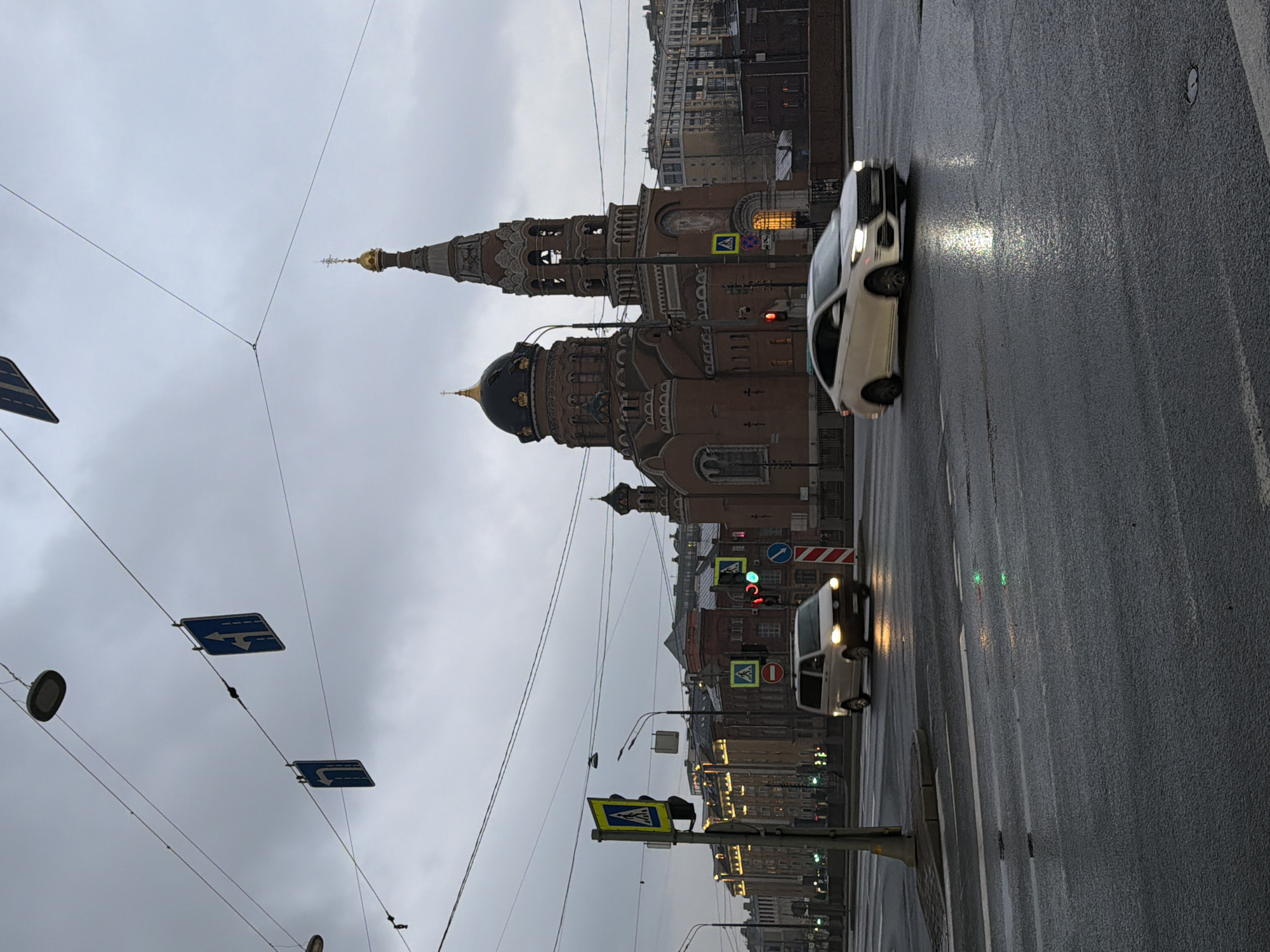 The image depicts a street scene in Saint Petersburg, Russia, on a cloudy day. The wet road surface suggests it has recently rained. Prominently featured on the left is the Church of the Assumption of the Blessed Virgin Mary, known for its distinctive onion domes and brick facade. Overhead, tram lines and various traffic signs are visible. A white car is moving along the wet road on the right-hand side of the street. Further down the street, more cars, traffic lights, and buildings can be seen.