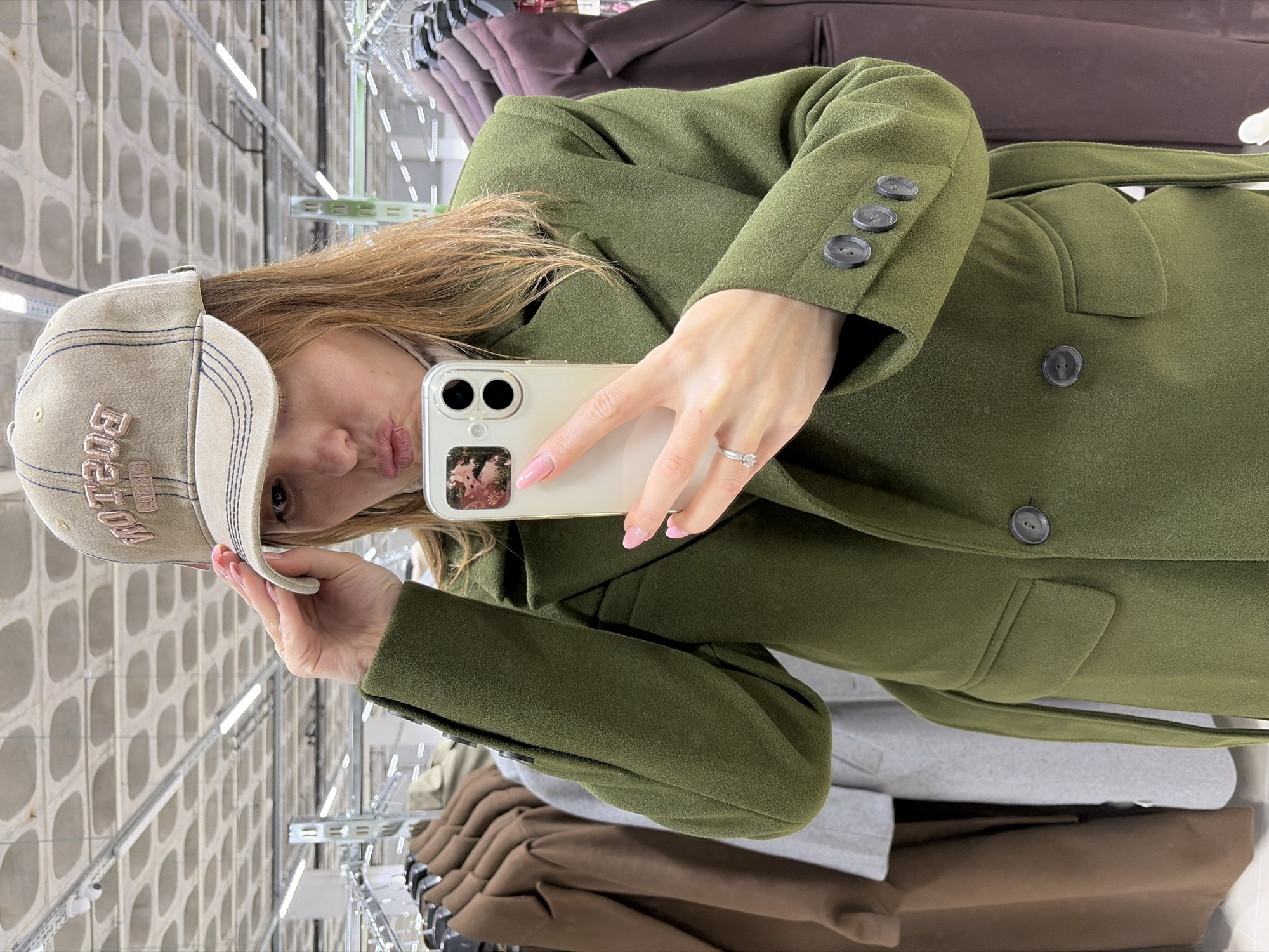 A young woman is captured taking a selfie inside what appears to be a clothing store in Izhevsk, Russia. The image is oriented vertically, with her head to the left and body extending to the right. She holds a light-colored smartphone with both hands, positioned in front of her face, with a slight pout on her lips as she looks towards the camera.
She wears a long, olive green coat with a textured fabric. The coat features three dark, circular buttons on the cuff of her right sleeve and one larger dark button visible on the front, near her waist. A matching fabric belt is tied around the coat. On her head, she sports a light-colored baseball cap with prominent blue stitching and raised pink lettering that clearly spells "BOSTON" on the front. Her nails are painted a light pink, and she wears a silver-toned ring with a small stone on her left ring finger. The smartphone is enclosed in a clear case, and a small picture or decal is visible on its back.
The background reveals a retail setting with clothing racks displaying other garments, including coats in shades of brown and grey. A distinctive concrete-like wall with a grid of square recesses is visible behind the racks, indicating an indoor location. Overhead lighting illuminates the scene. The overall activity depicts a casual moment of trying on clothes and capturing a selfie, likely during a shopping outing in a contemporary store environment.